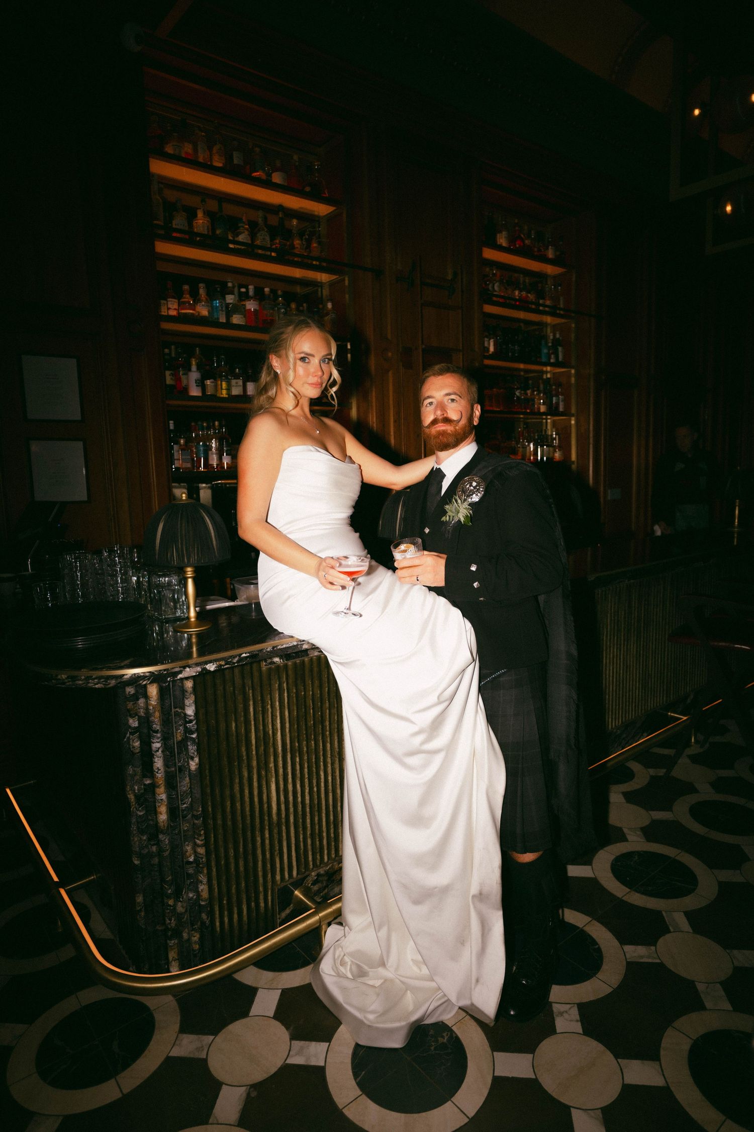 A couple dances together at a dimly lit bar venue with wooden shelves of bottles in the background.