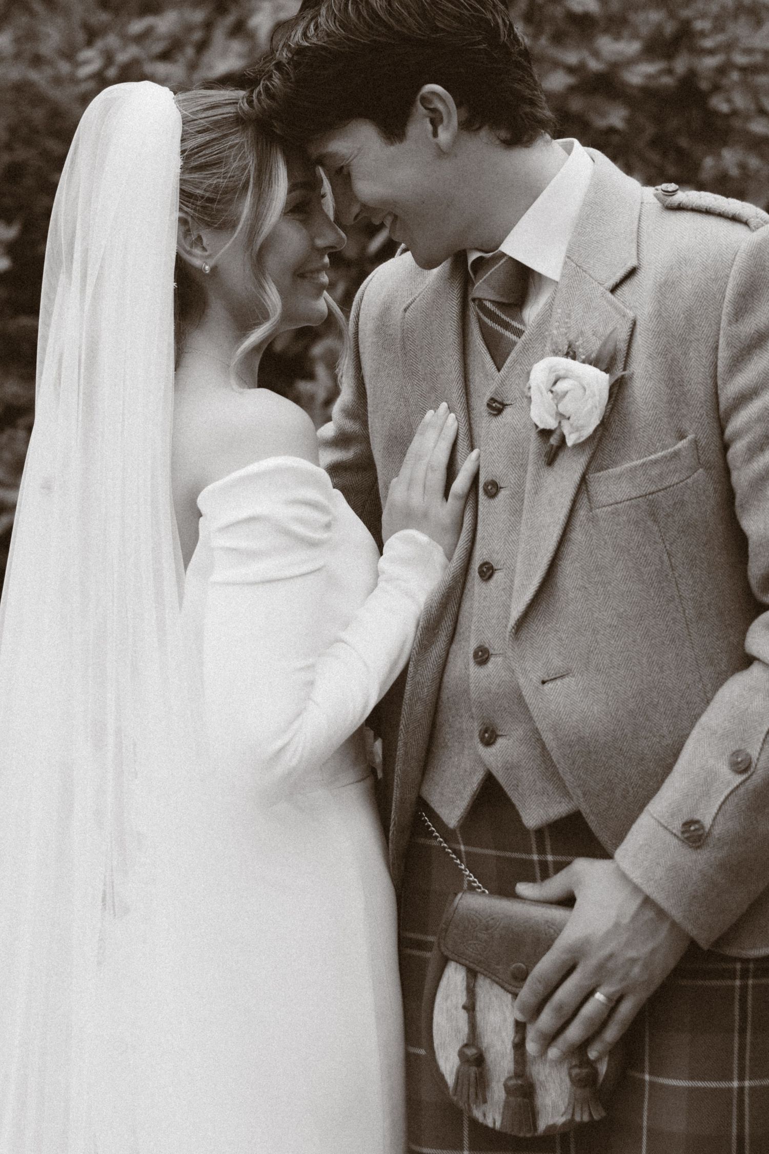 Black and white photo of a bride in white gown and veil with groom in Scottish kilt suit sharing an intimate moment.
