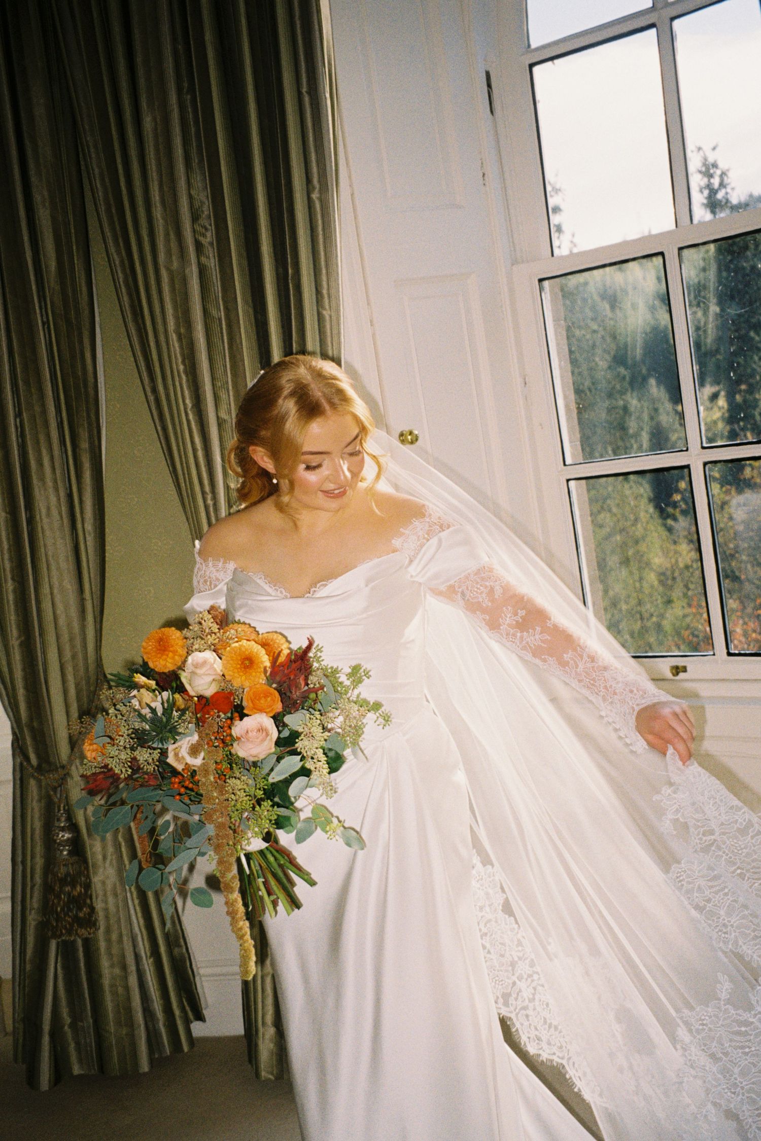 A bride in a flowing white lace wedding dress twirls near large windows with green curtains and holds a colorful bouquet.