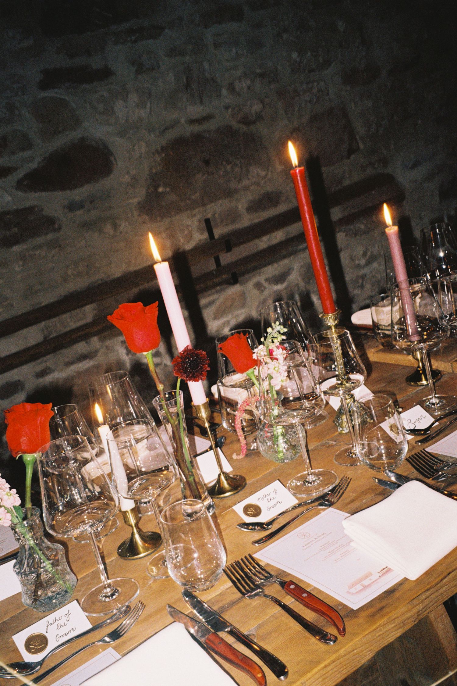 An elegant candlelit dinner table set with wine glasses, white candles, and red tulips in a rustic stone cellar setting.
