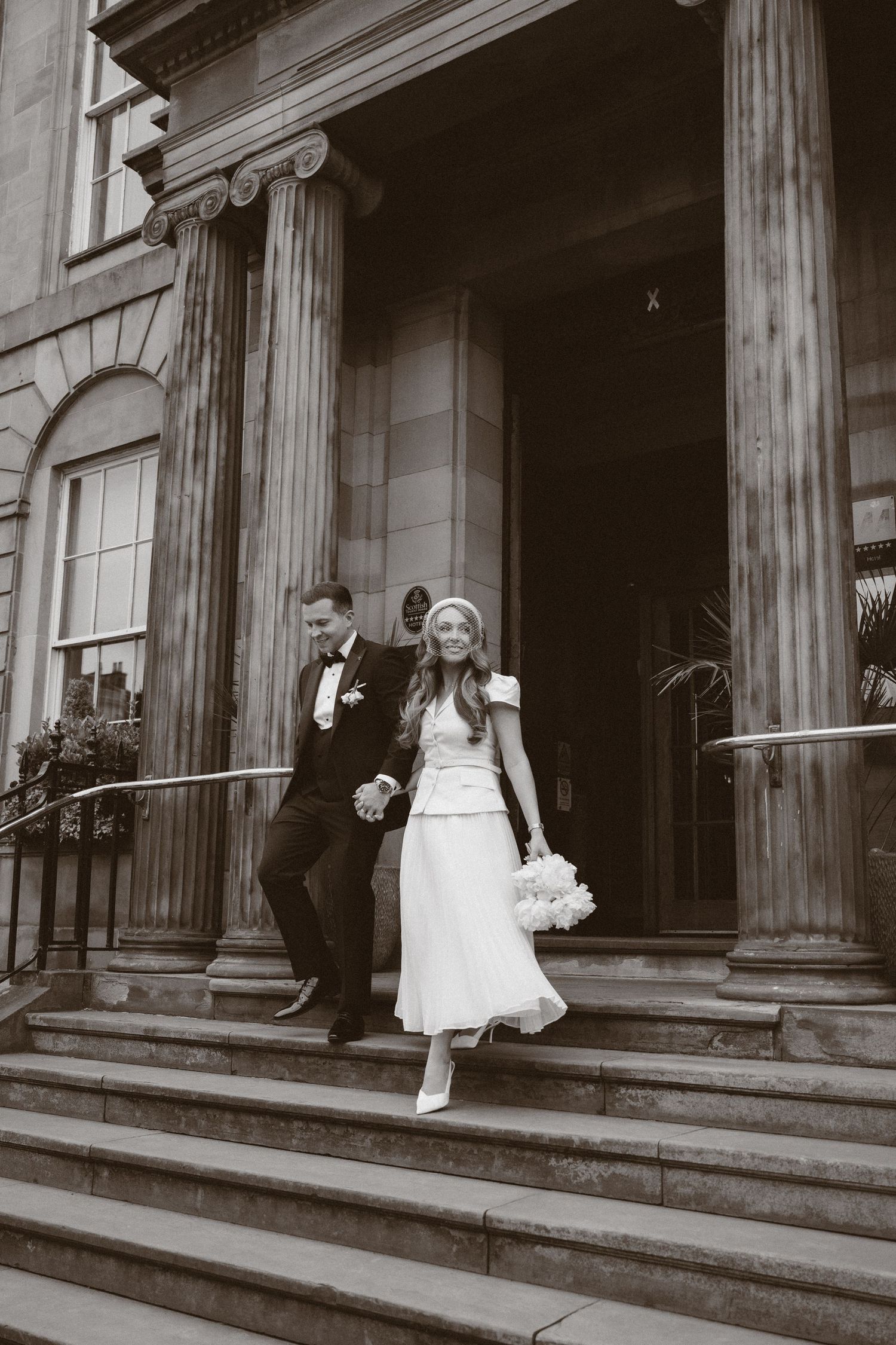 Black and white photo of a bride and groom descending stone steps of a grand building with columns on their wedding day.