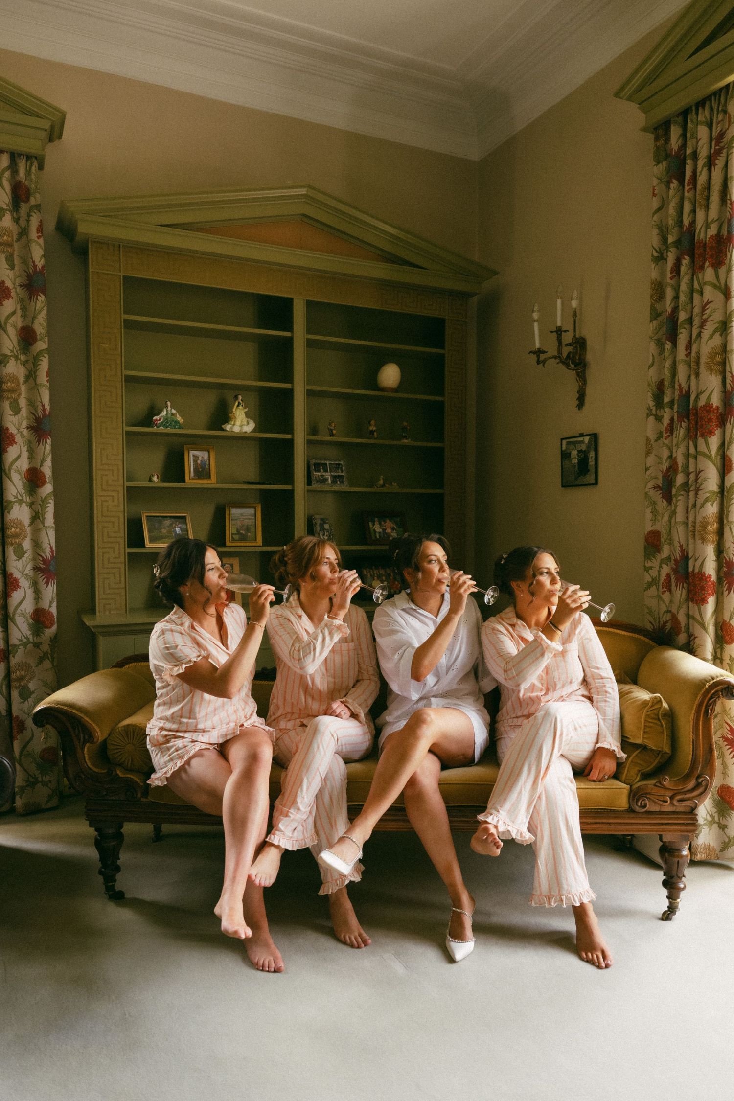 Four people in light pink robes and nightwear sit together laughing on an elegant vintage sofa in a classic interior setting.