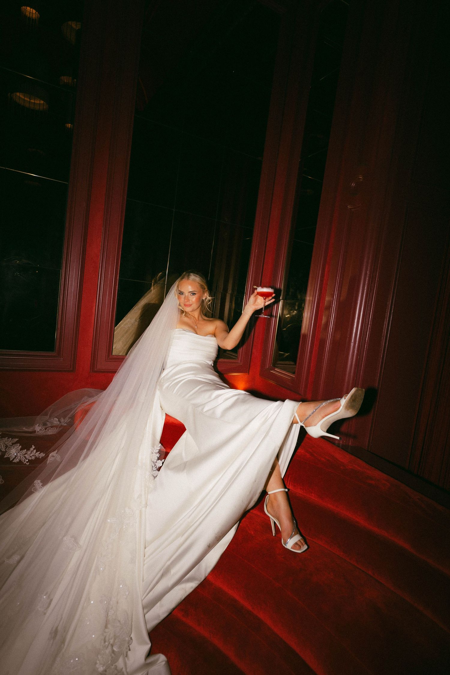 A flowing white wedding dress creates a dramatic silhouette against red carpeted stairs in a dark room.