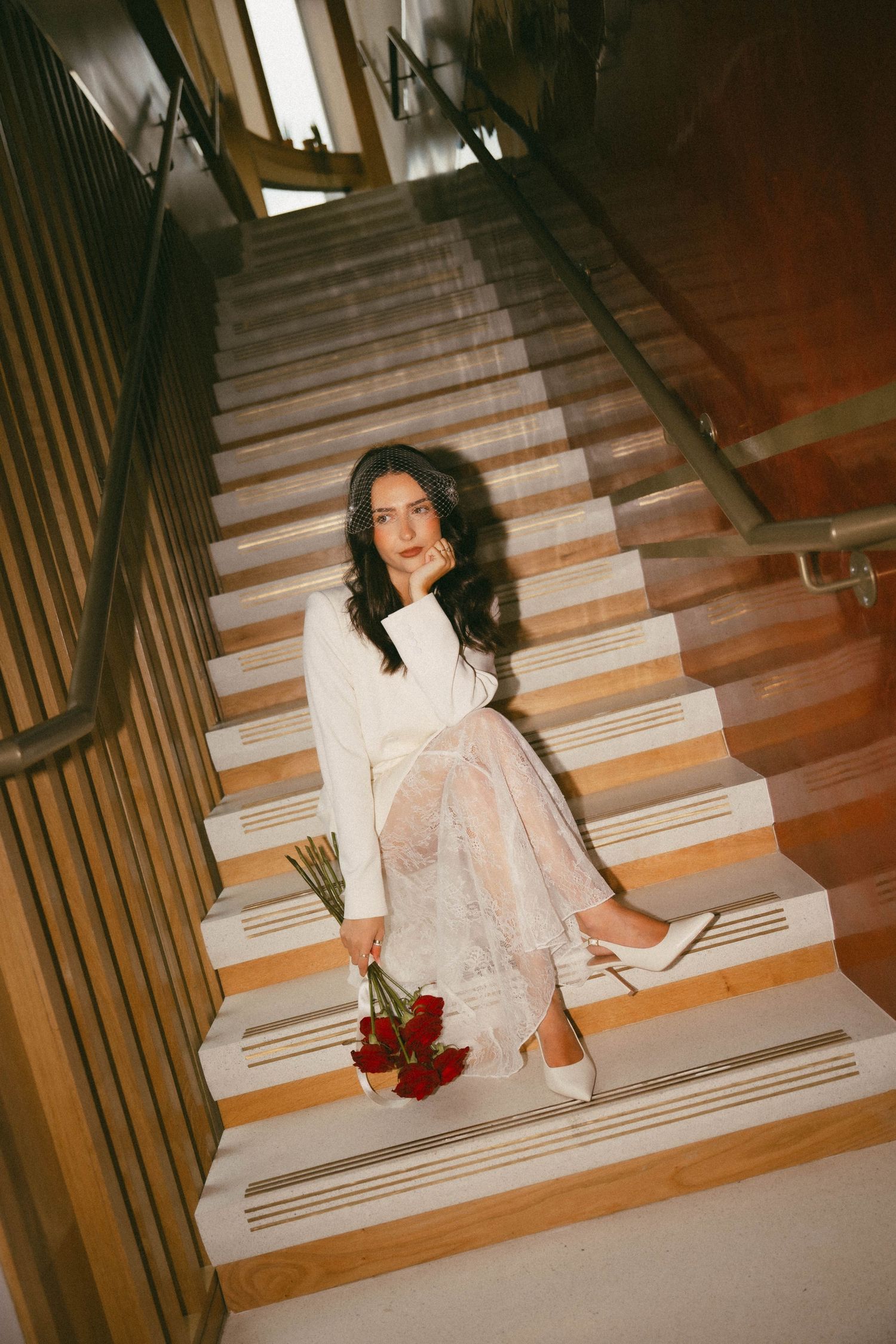 A person in a white dress sits on wooden stairs holding red flowers in a vintage-style indoor photograph.