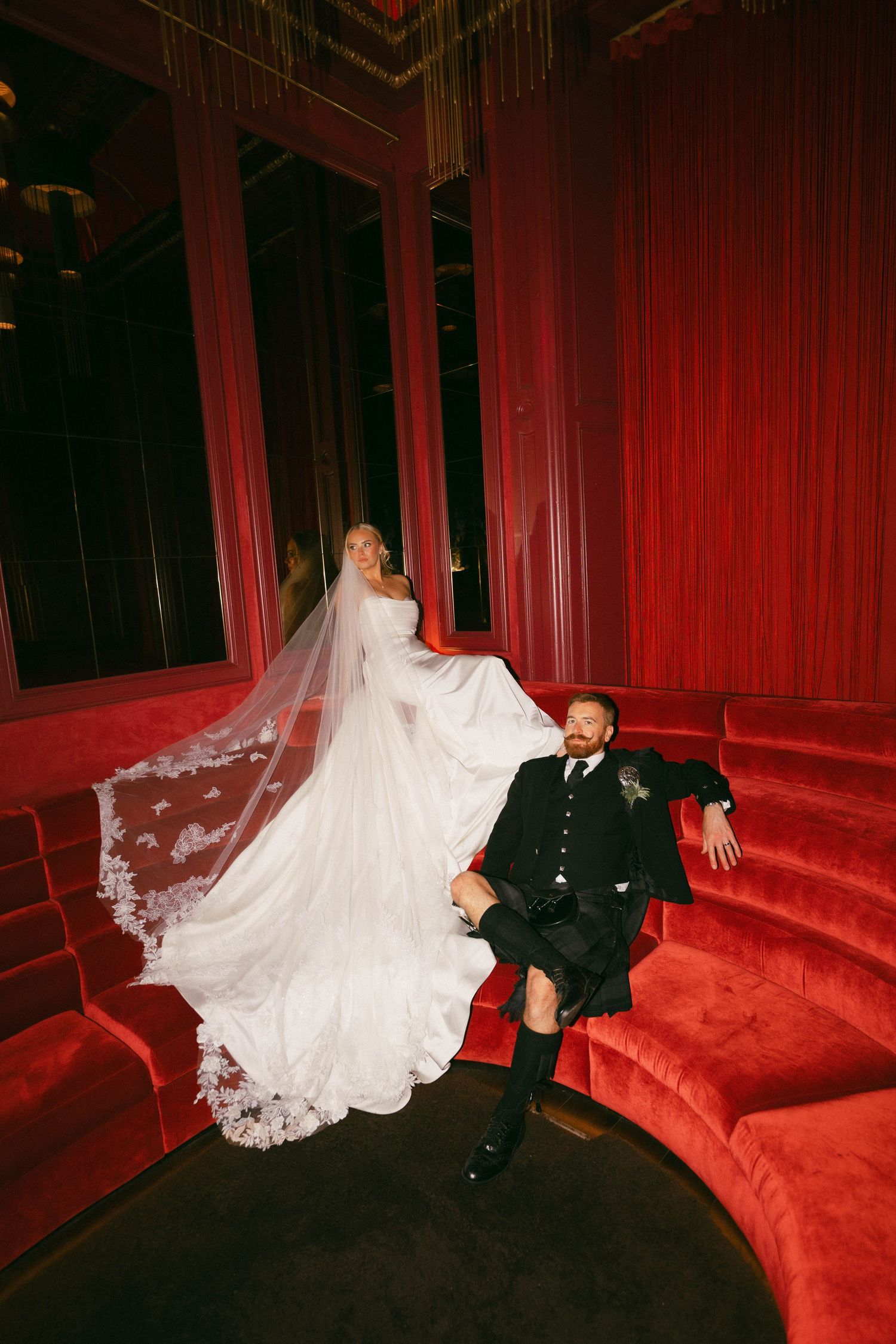 A couple in wedding attire poses dramatically on a plush red velvet couch in a dimly lit room.