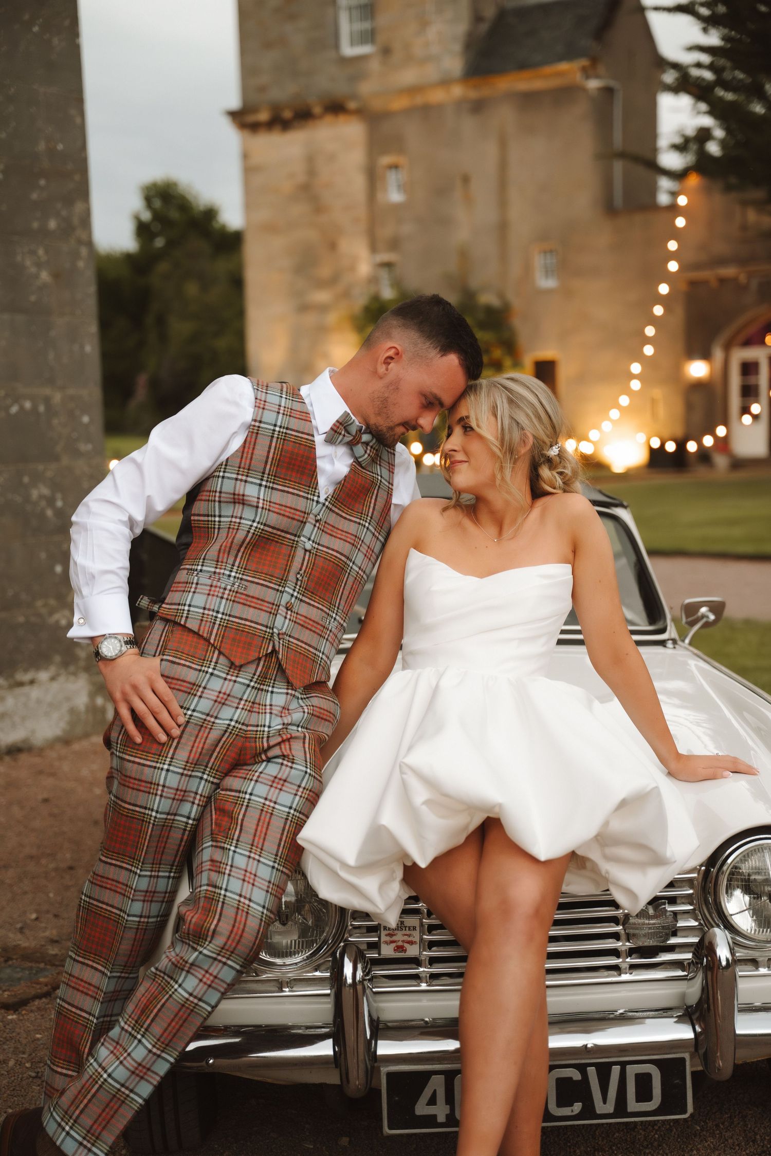 Bride in short white dress and groom in tartan suit lean against vintage car outside a Scottish castle at dusk.