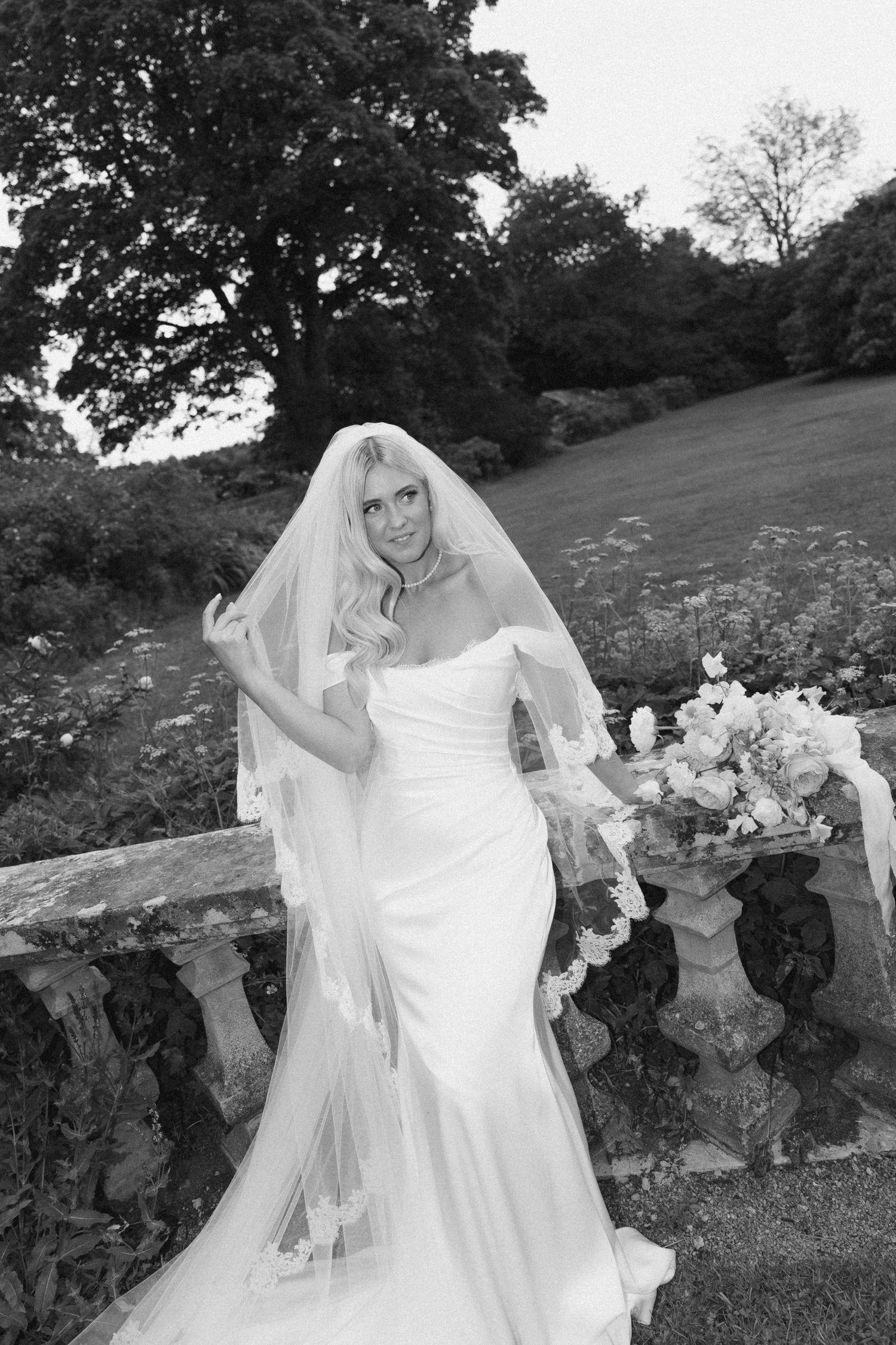 A bride in a white mermaid wedding dress and long veil poses on a stone wall in a garden setting.