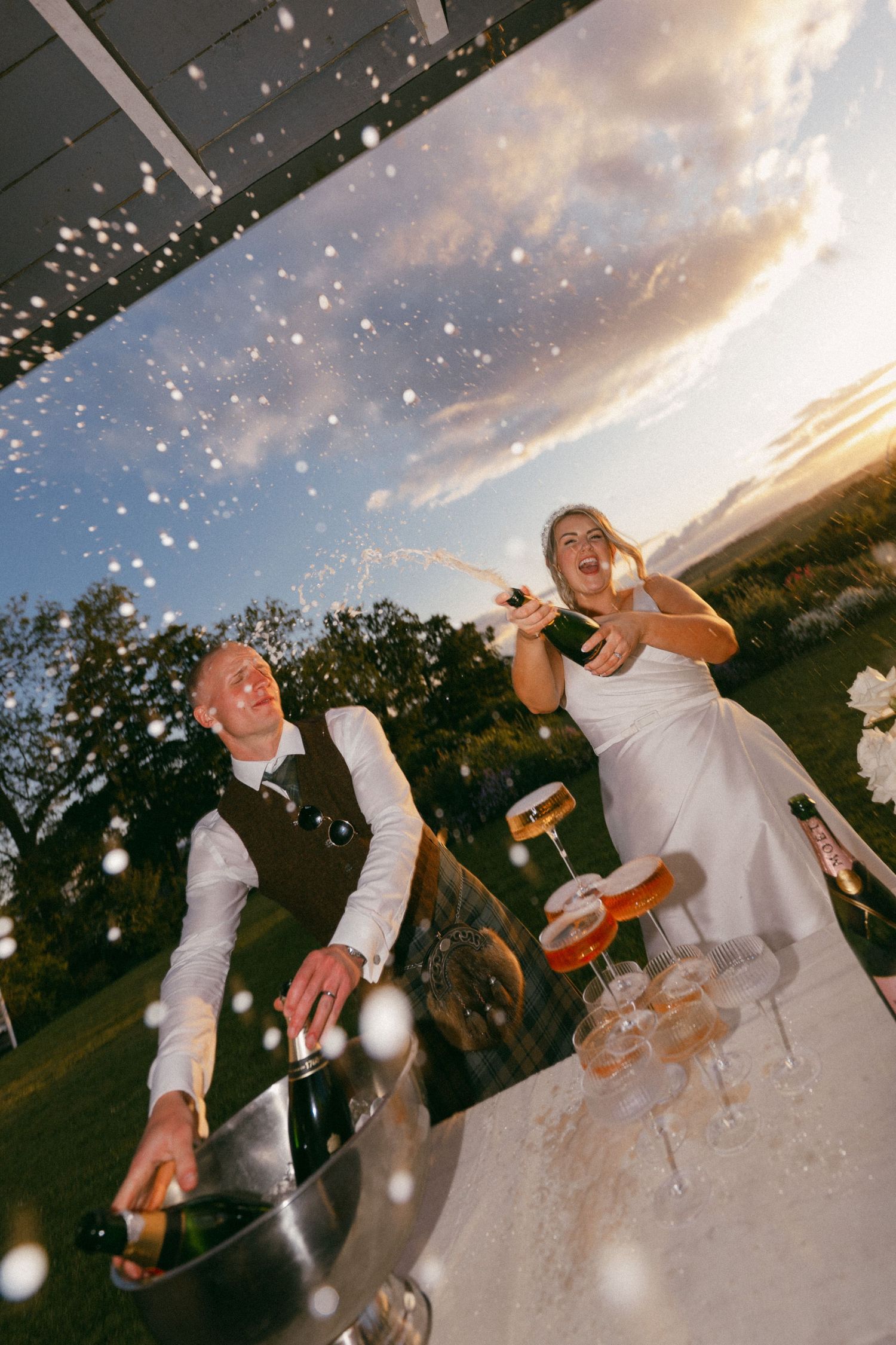 A joyful couple pops champagne bottles on their wedding day as bubbles float through the golden sunset sky.