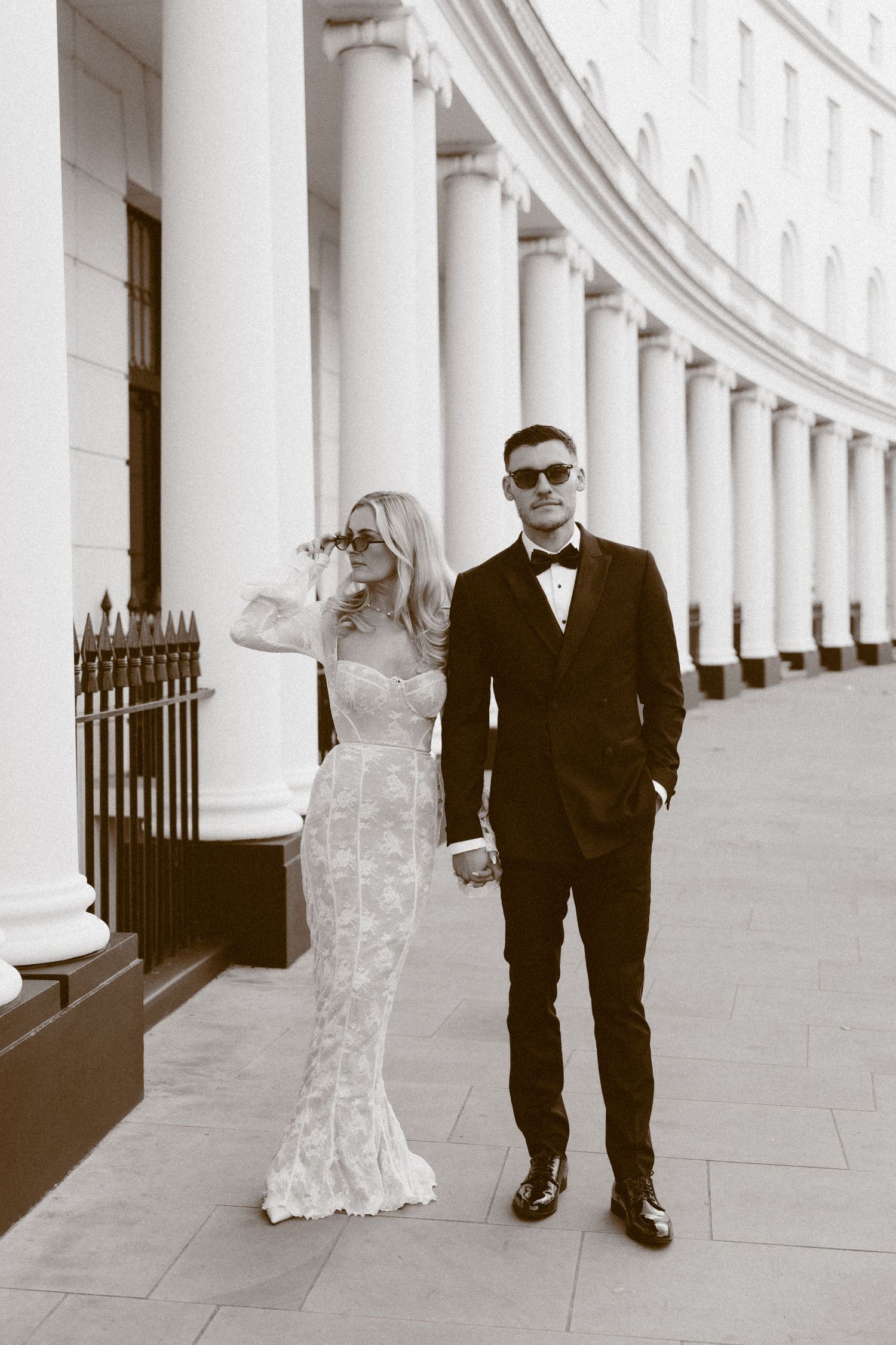 A bride in a lace wedding dress and groom in black suit pose together along a white columned walkway in black and white.