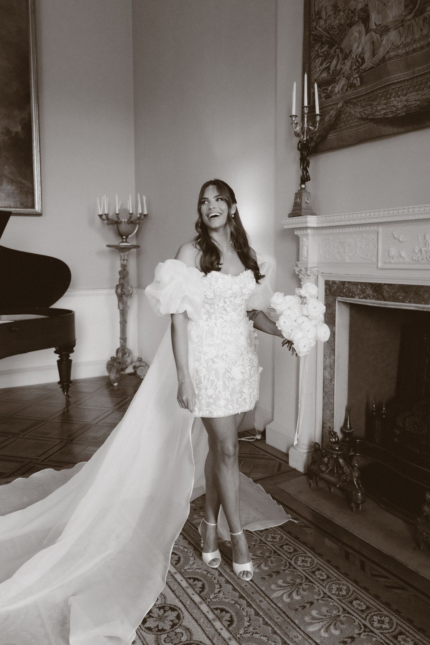 A bride in a short white off-shoulder wedding dress with a long train poses elegantly by a fireplace.