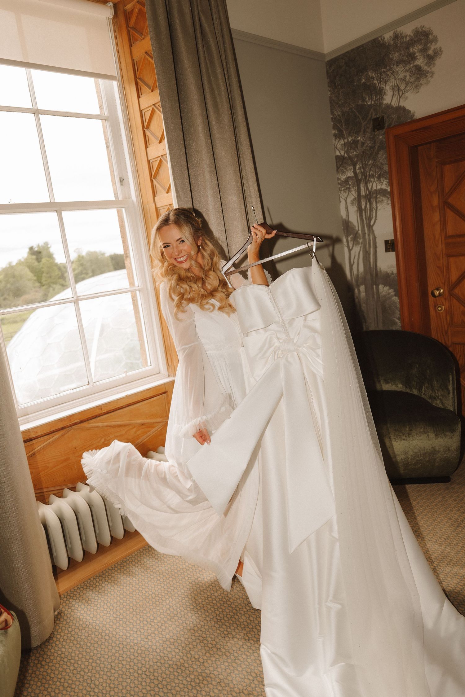 Bride in white wedding dress holding gown on hanger near sunlit window in elegant room.