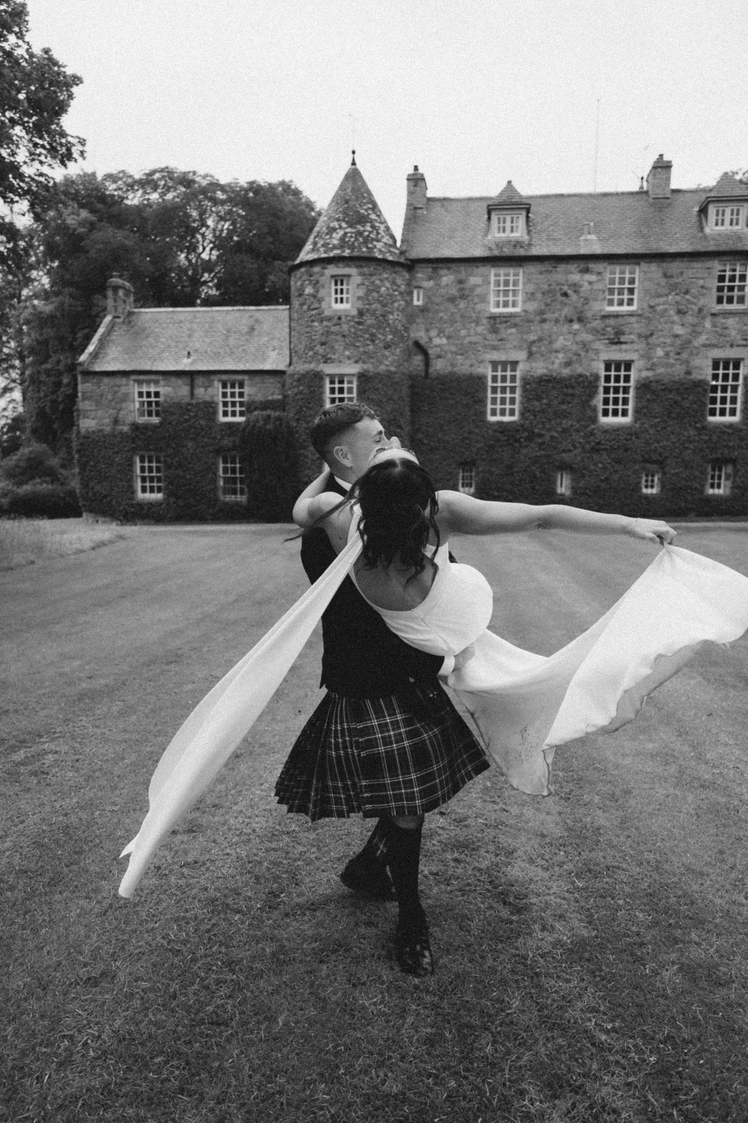 A dancer in a kilt spins gracefully with flowing fabric on the lawn of a historic stone castle in black and white.