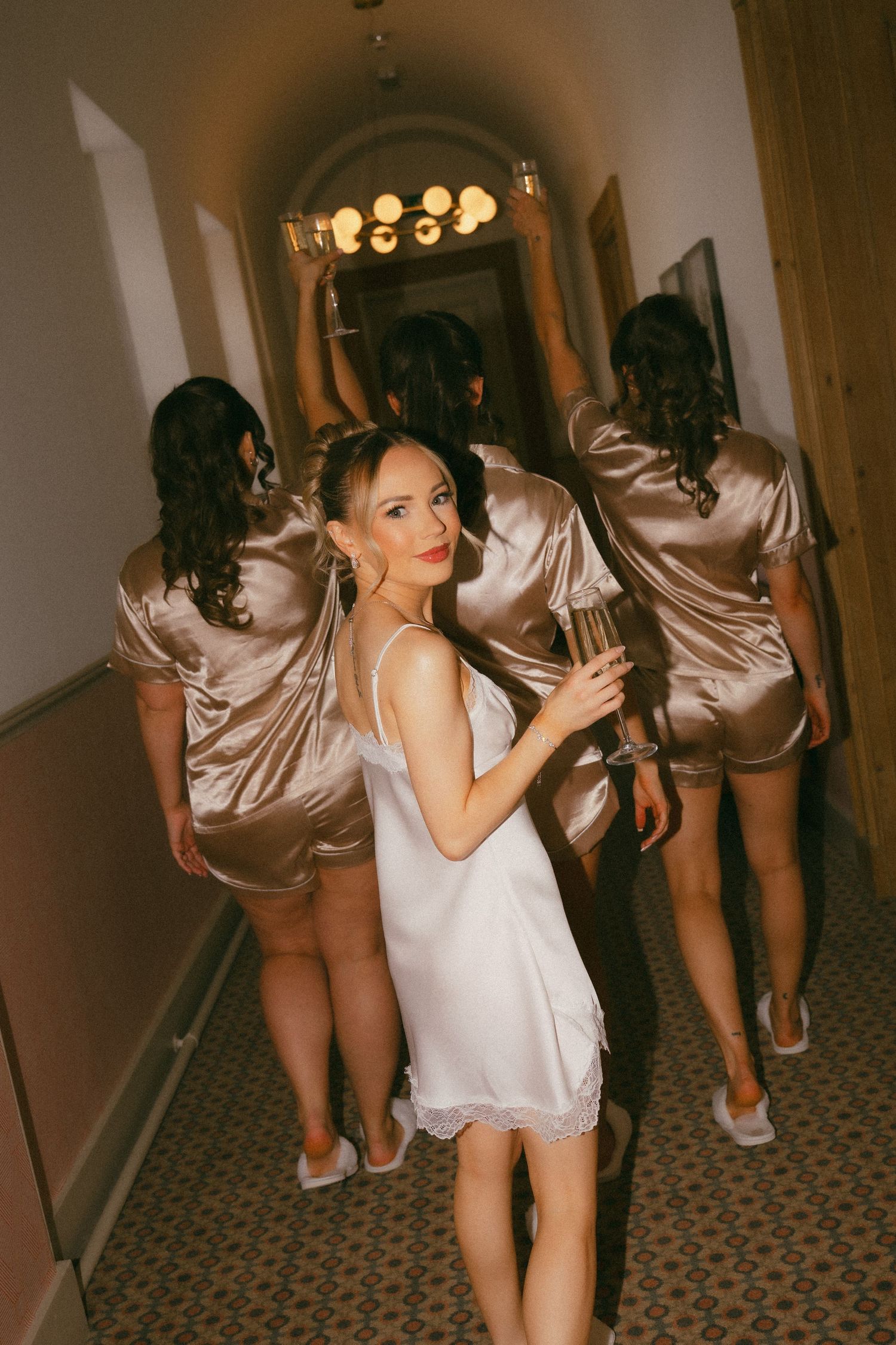 Bride in white slip dress holds champagne with bridesmaids in gold satin pajamas walking down hotel hallway.