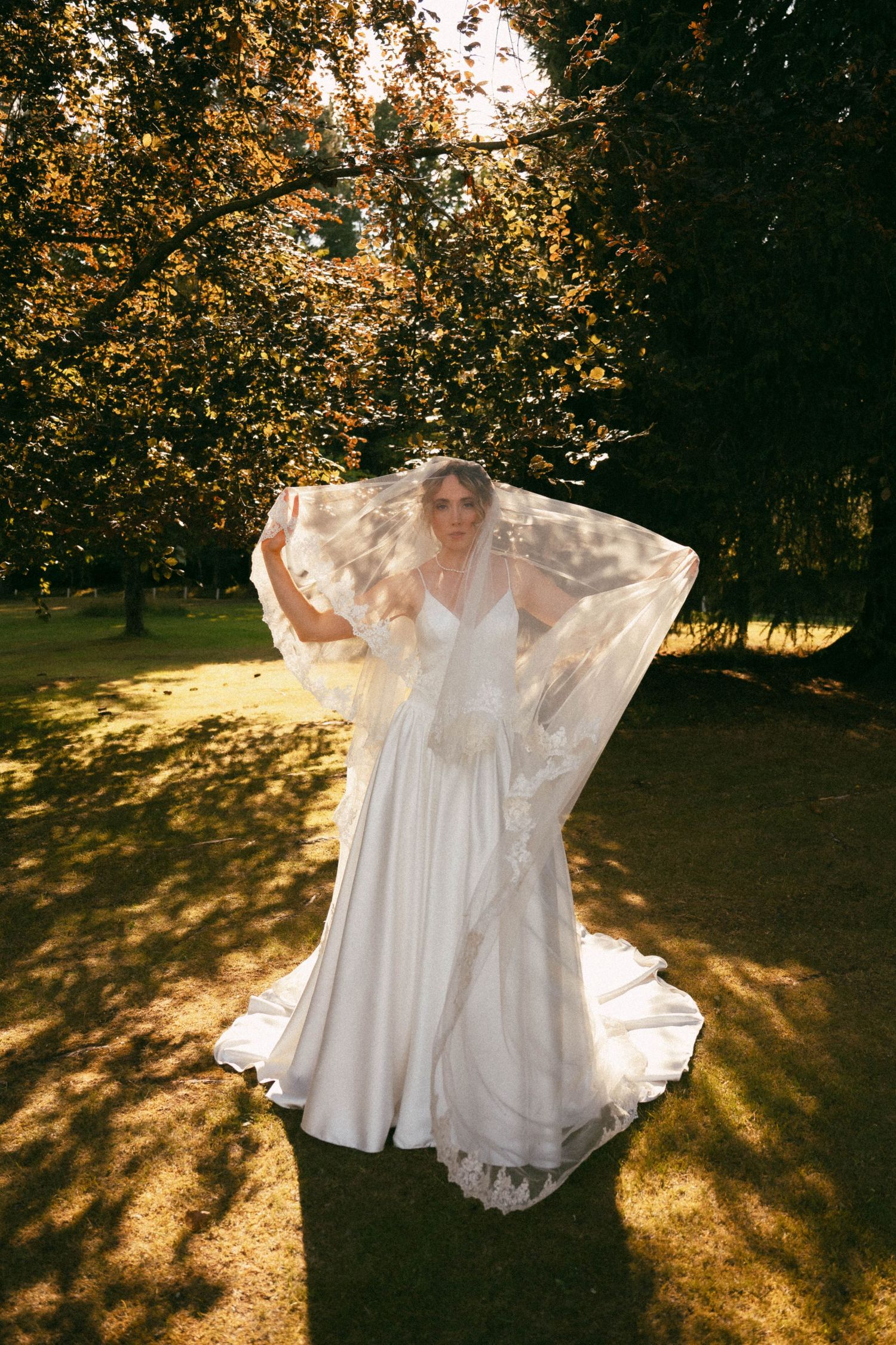 White wedding dress twirls in golden sunlight casting dreamy shadows across a grassy field at sunset.