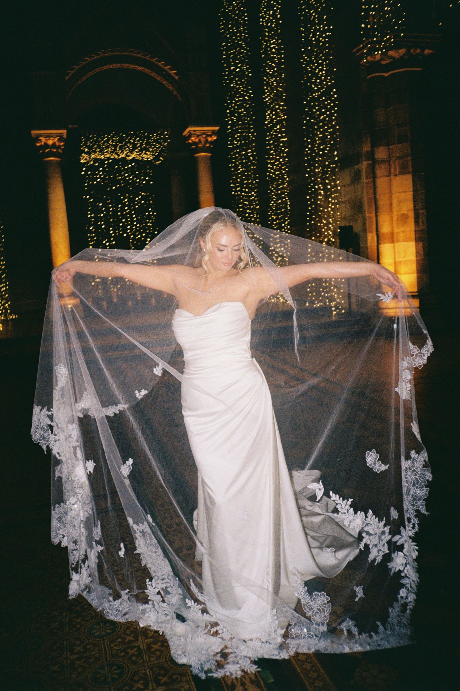 A bride in a white dress poses with an ornate lace veil against a backdrop of twinkling lights at night.