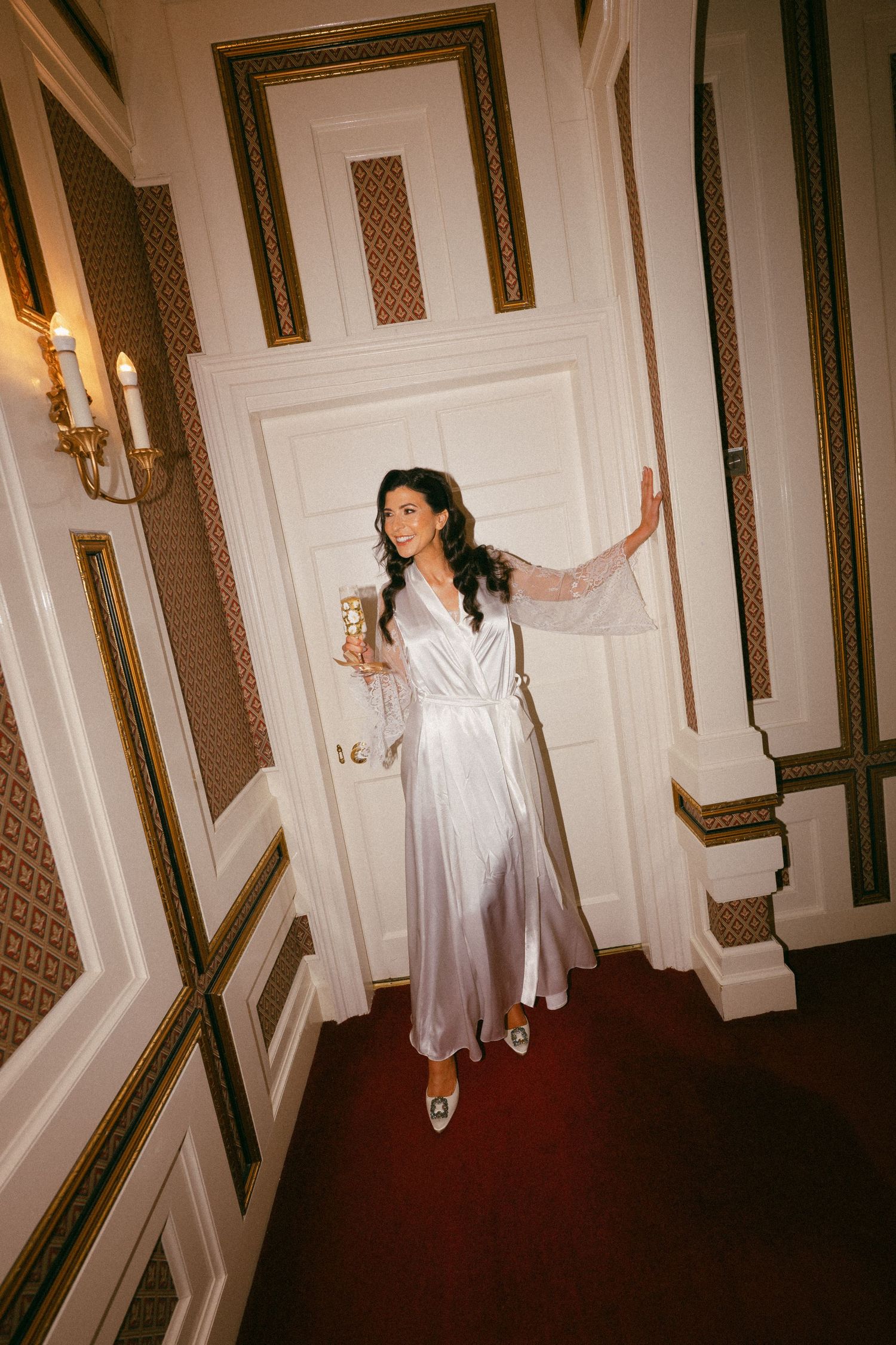 Figure in white silk robe walks through ornate hallway holding champagne glass.