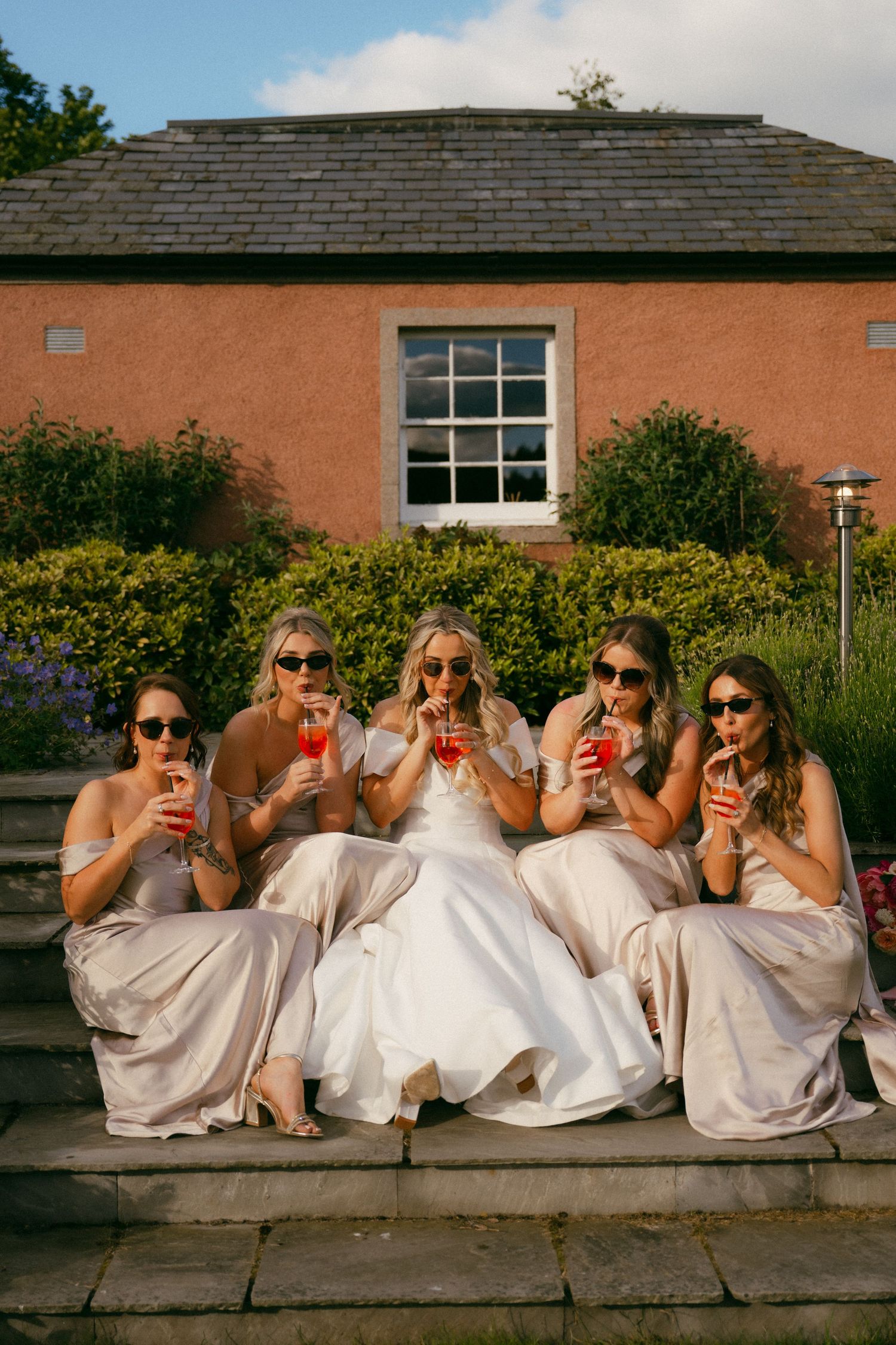 Bride and four bridesmaids in champagne dresses sipping cocktails on stone steps outside a pink brick building.