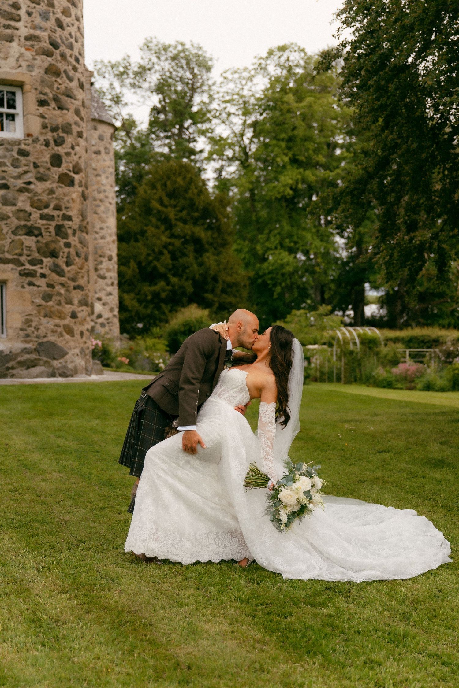 Bride and groom share a romantic kiss on green lawn beside a Scottish stone castle, bride holding white bouquet.