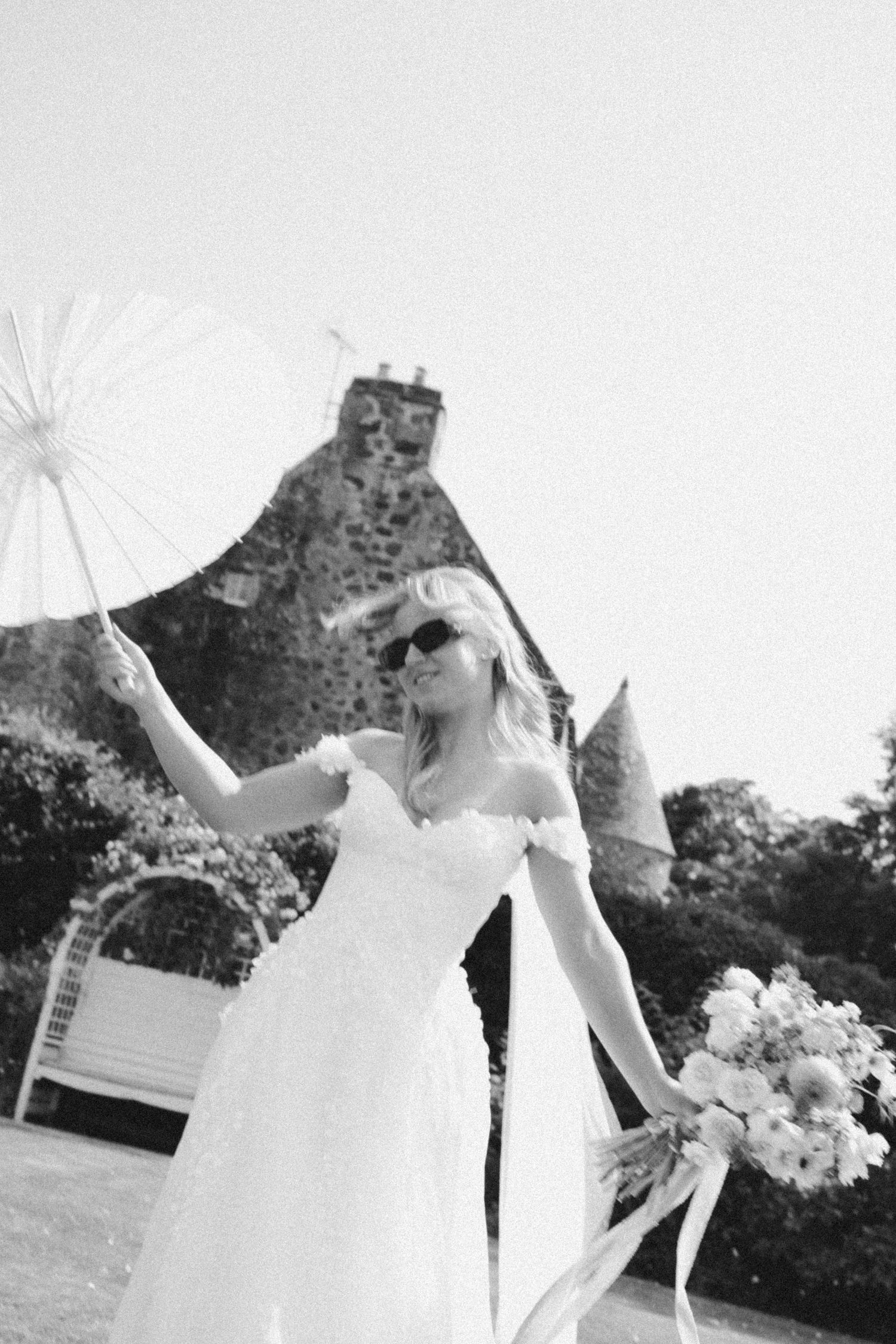 A bride in a white off-shoulder wedding dress twirls while holding a parasol outdoors in a garden setting.