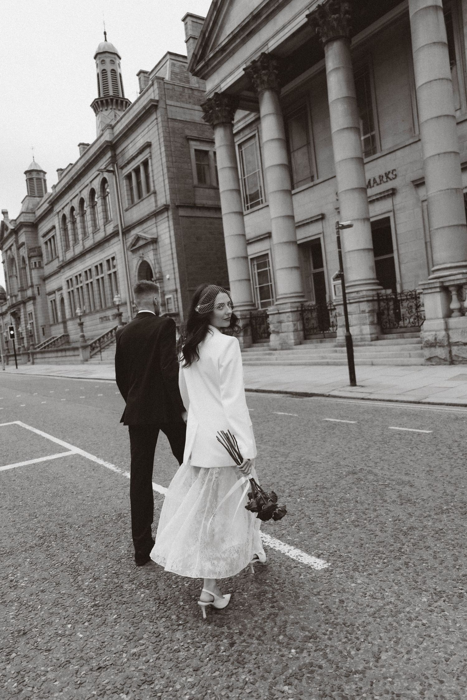 A wedding couple walks together on cobblestone streets alongside classic stone architecture with white columns.