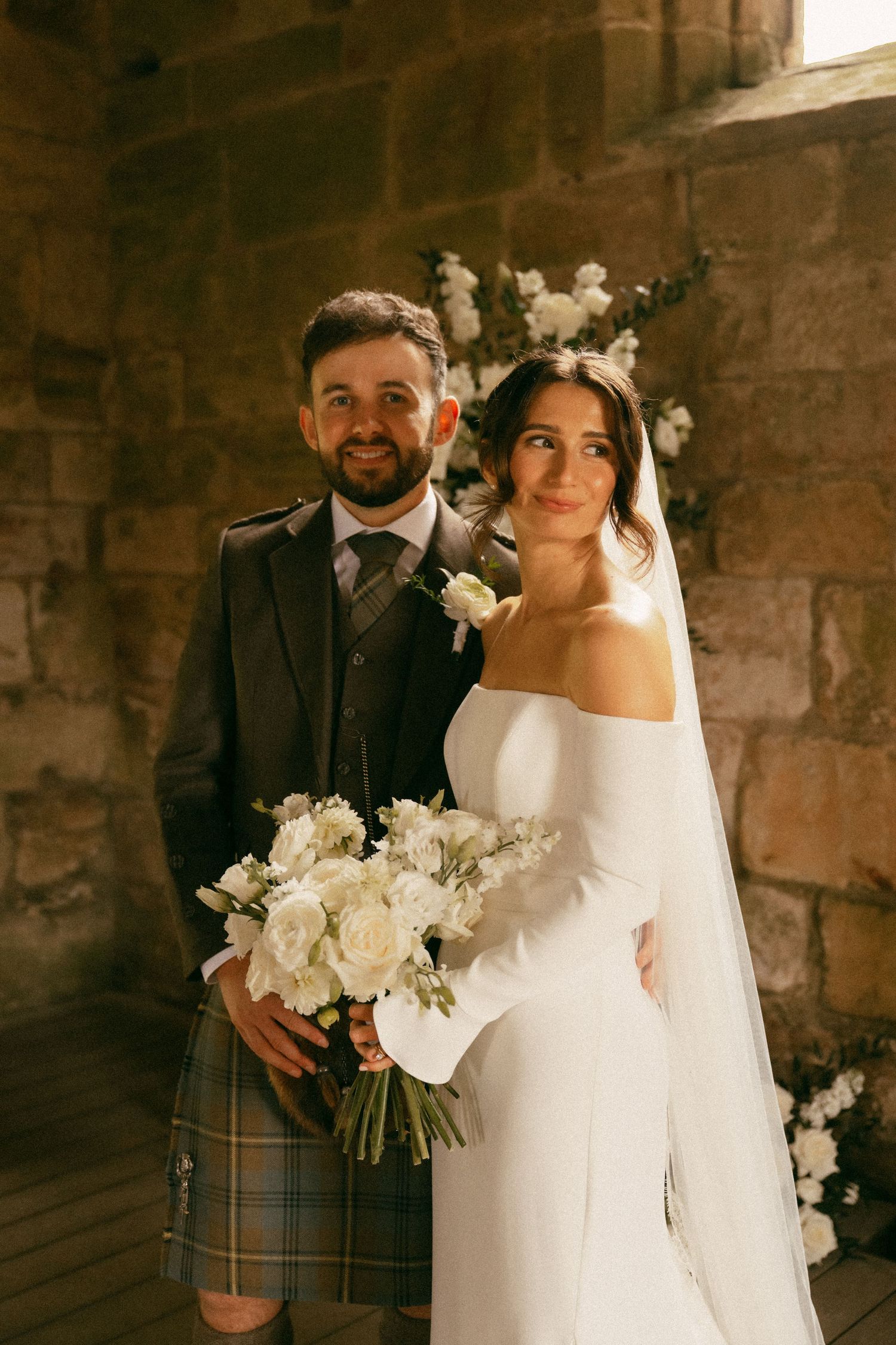 Bride in white off-shoulder gown with veil holds bouquet beside groom in Scottish tartan kilt inside stone venue.