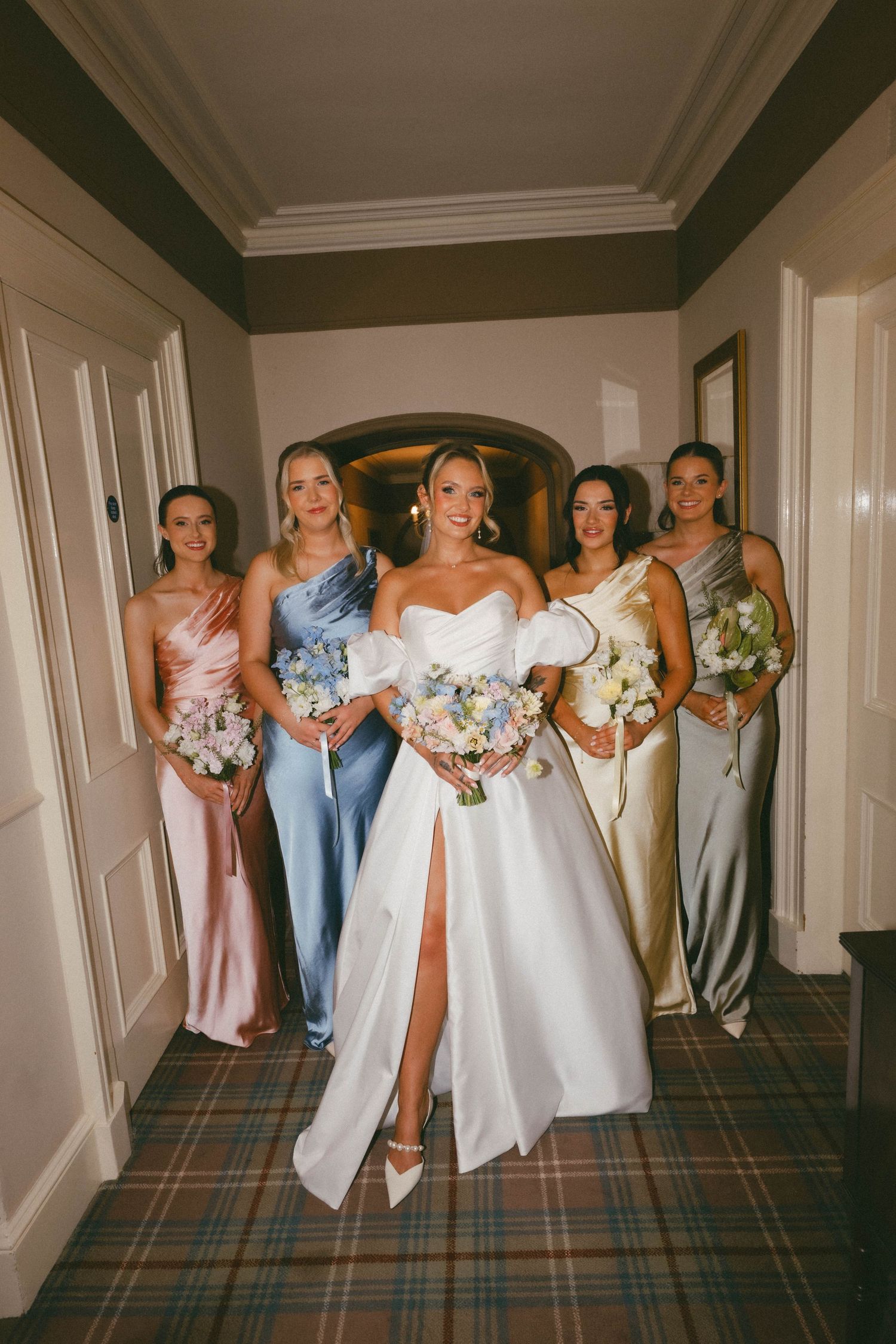 Bridesmaids in pastel and light blue dresses stand together in a hallway holding bouquets at a wedding venue.