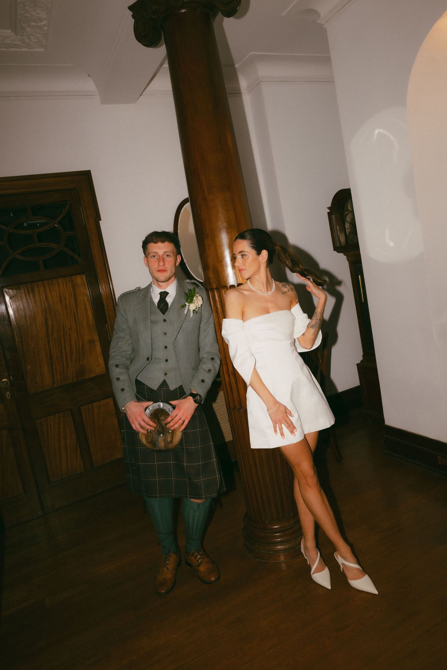 A couple in wedding attire sits together in a dimly lit room with wooden beams and white walls.