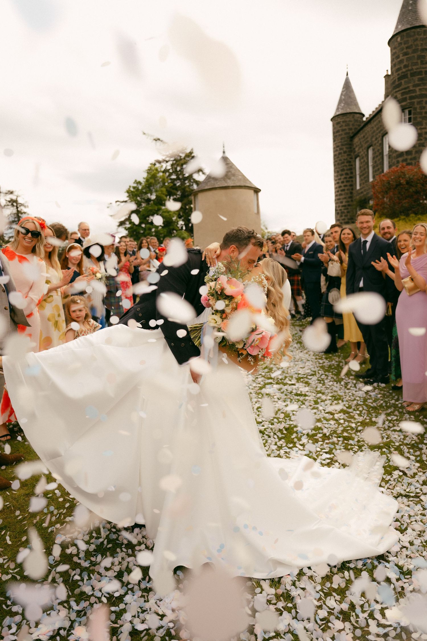Bride and groom share a romantic kiss surrounded by guests throwing white confetti outside a stone castle venue.