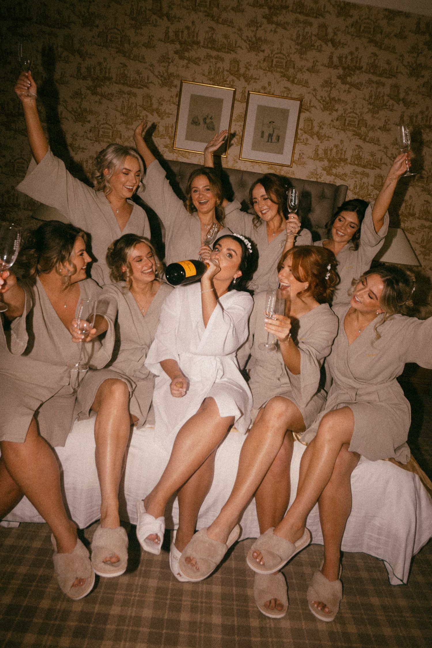 A group of bridesmaids in matching robes celebrate with champagne on a couch. Aberdeen wedding photographer, direct flash, meldrum house