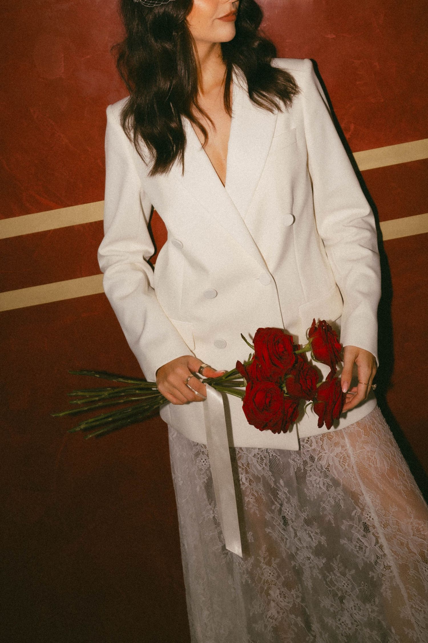 Person in white blazer holds deep red roses against a warm brown background.