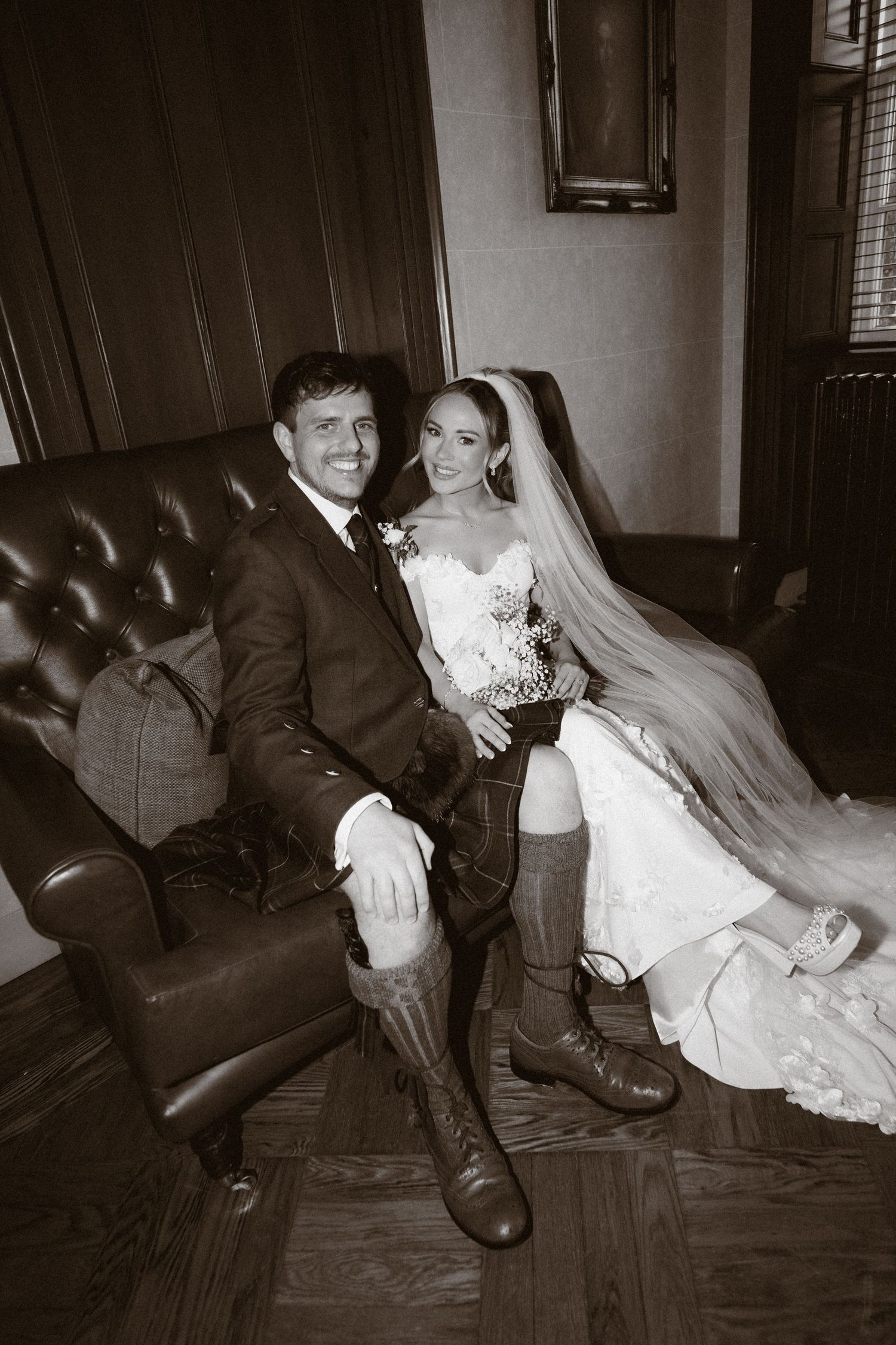 Bride and groom in wedding attire with boots sit together on leather chair in sepia-toned wedding portrait.
