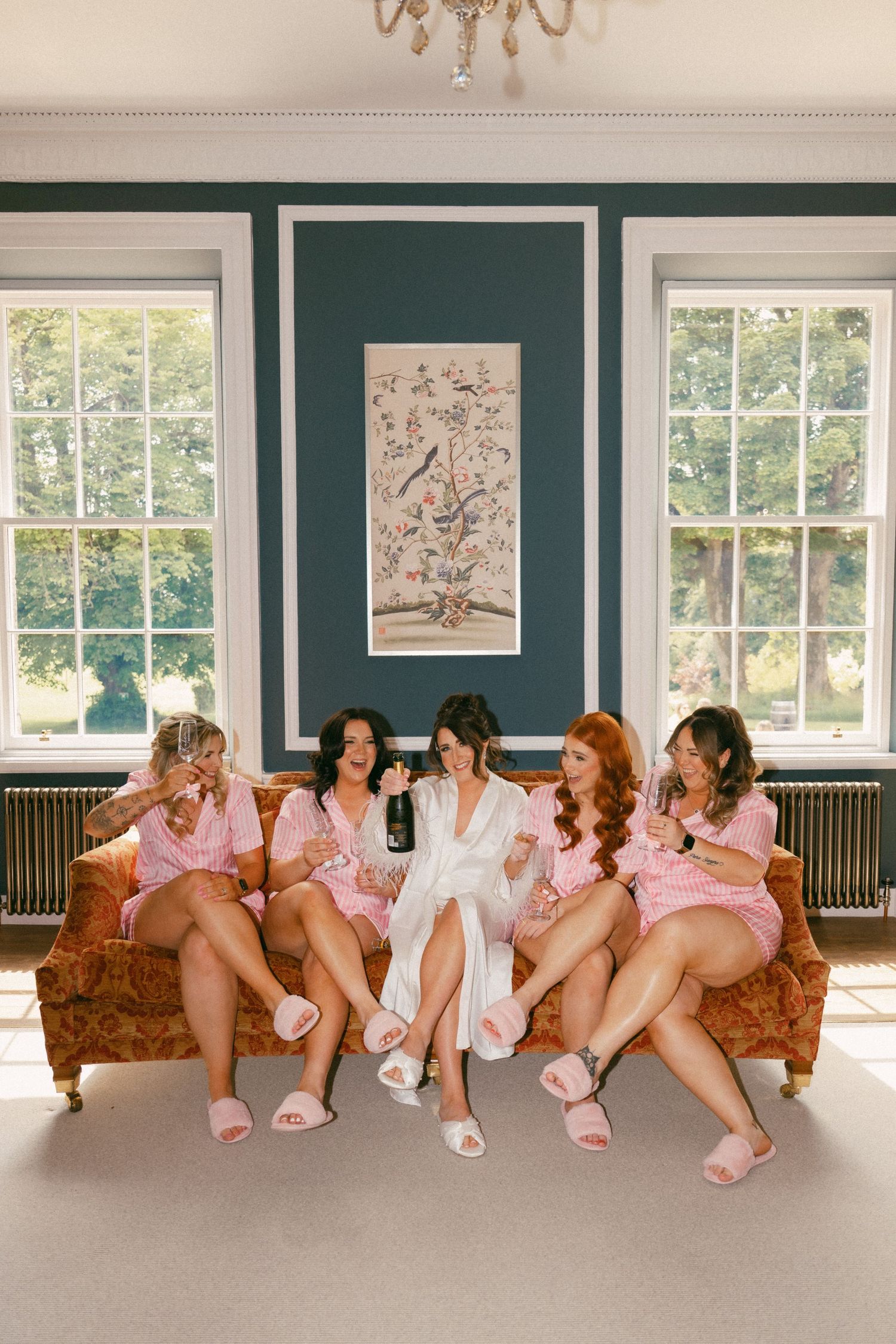A group in pink and white robes sits together on an elegant vintage sofa in a bright room with large windows.
