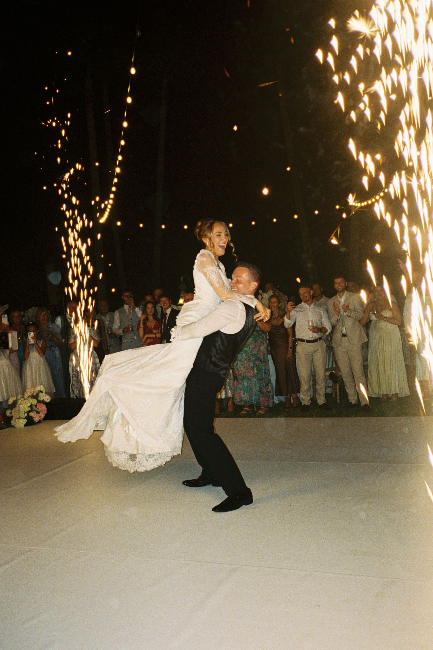 Groom lifts bride during first dance with sparkling fireworks at elegant outdoor wedding reception.