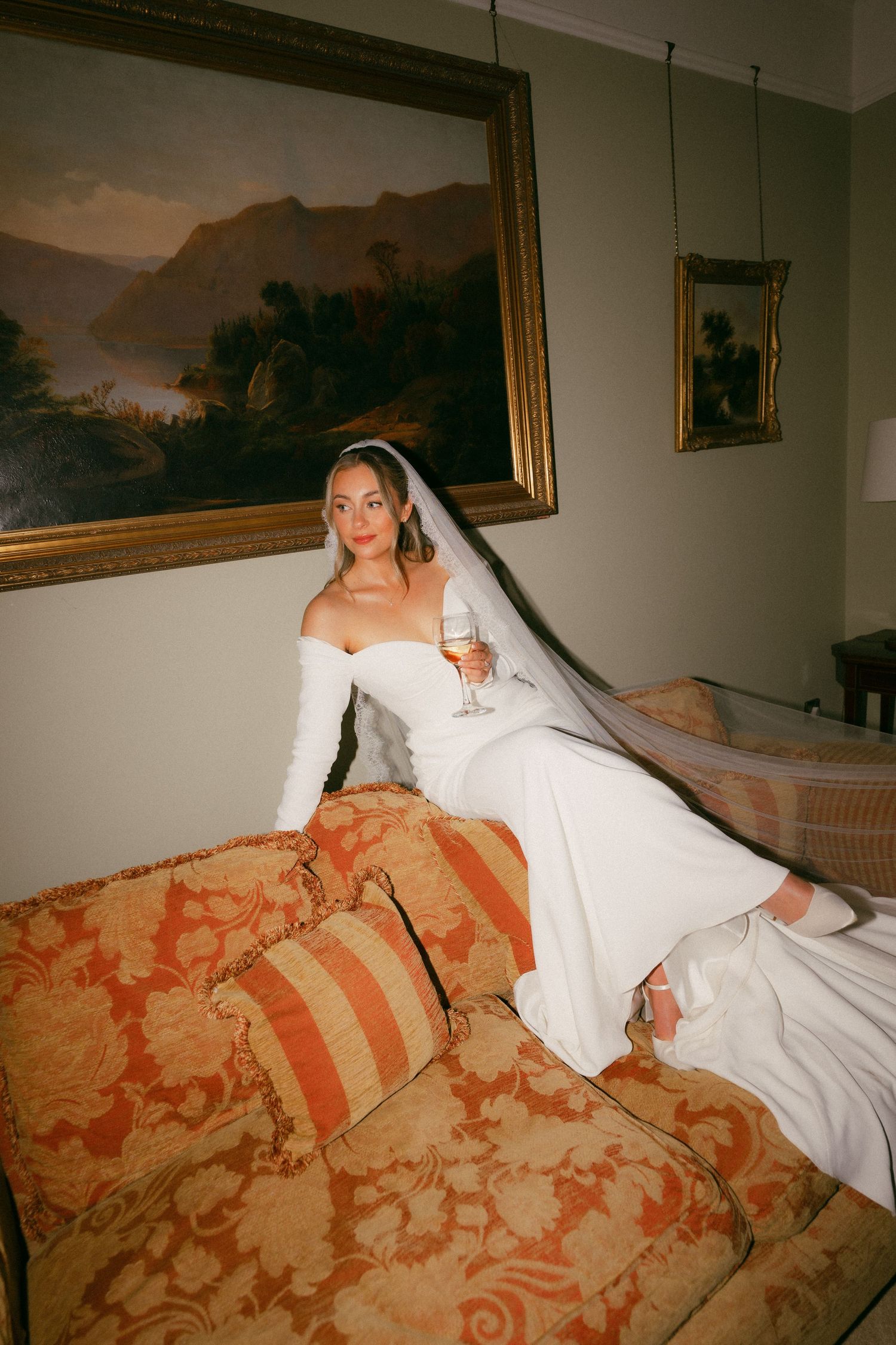 Bride in white off-shoulder gown and veil sits on ornate sofa holding champagne glass before classic oil painting.