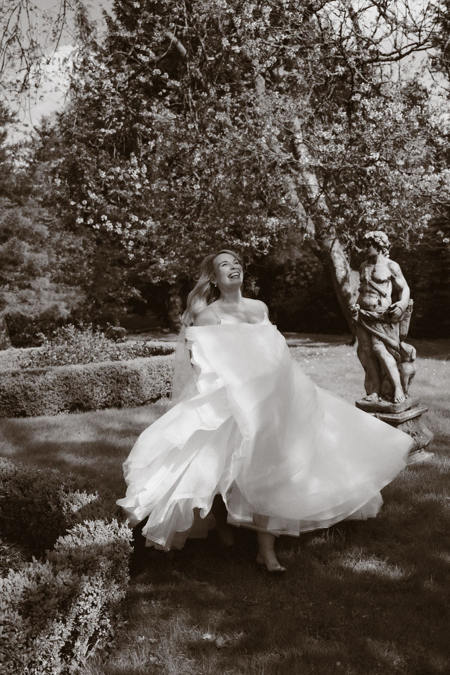 Black and white photo of a joyful bride in a flowing white gown twirling in a garden with a classical statue.