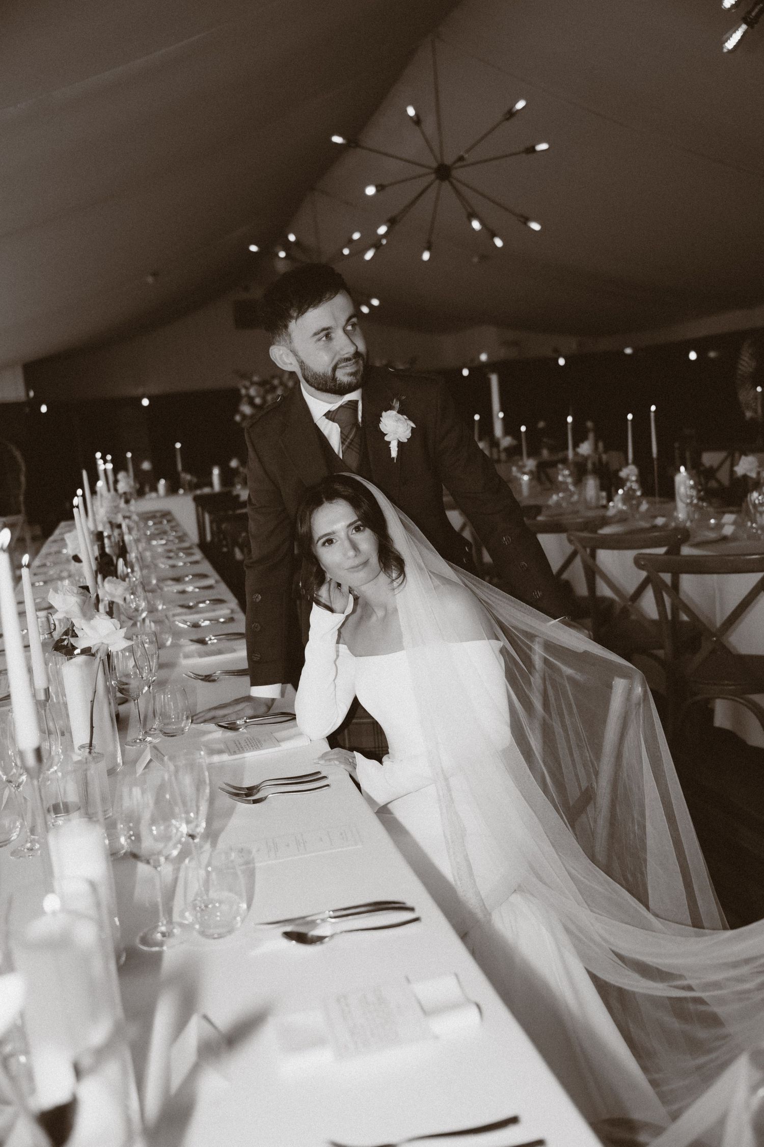Black and white photo of bride and groom at elegant wedding reception table with candles and fairy lights.