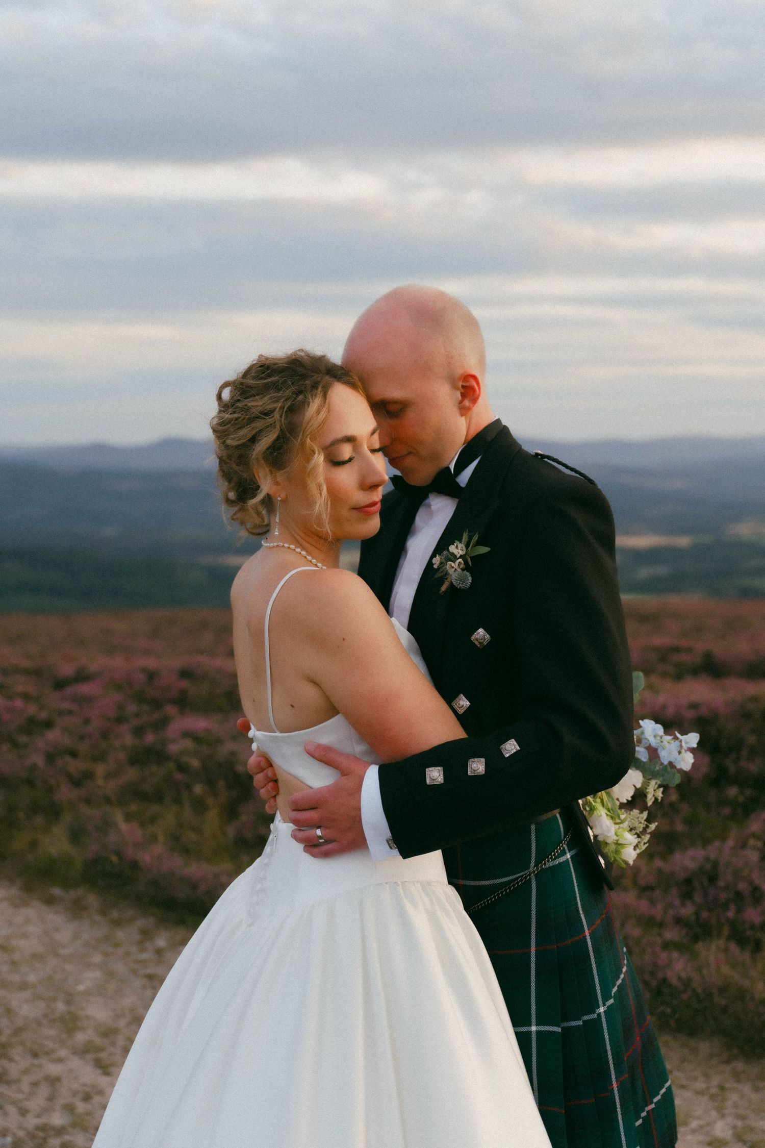 A couple embraces during their Scottish highland wedding, with the bride in white and groom in traditional kilt against mountains.