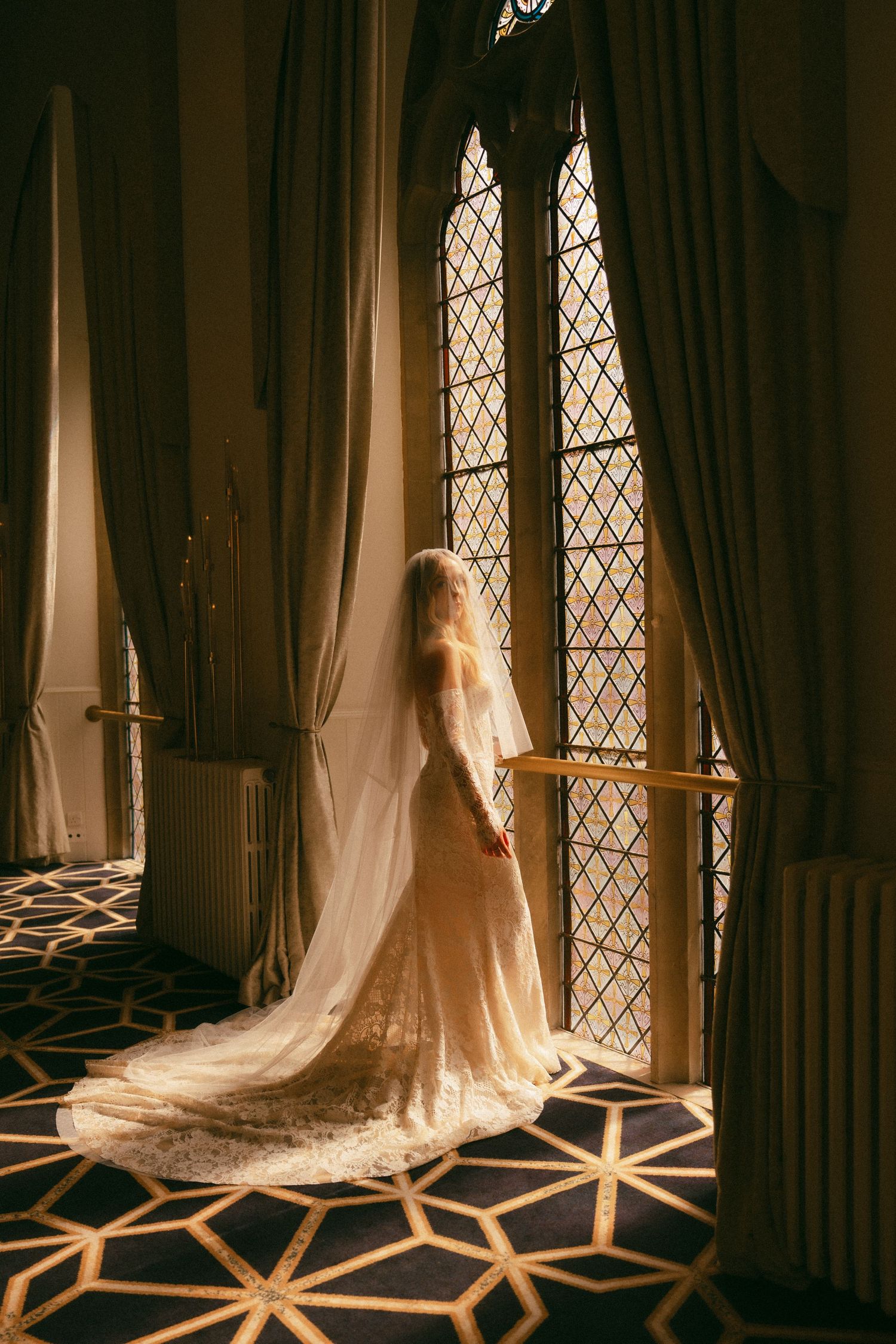 Bride in lace gown and long veil stands by ornate Gothic window, bathed in warm light on patterned carpet.