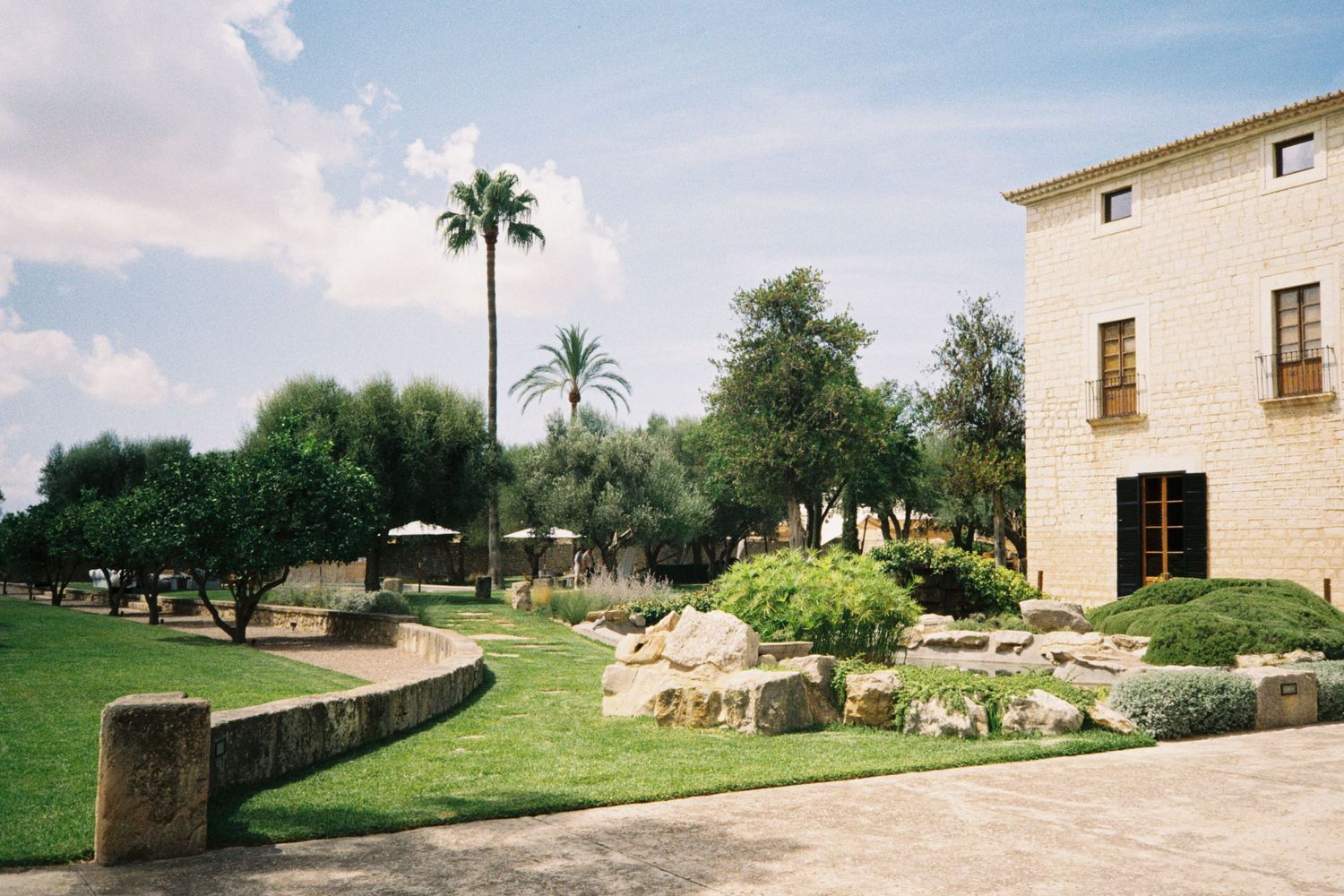 Lush Mediterranean garden with palm trees, stone landscaping, and a rustic stone building facade under a clear blue sky.
