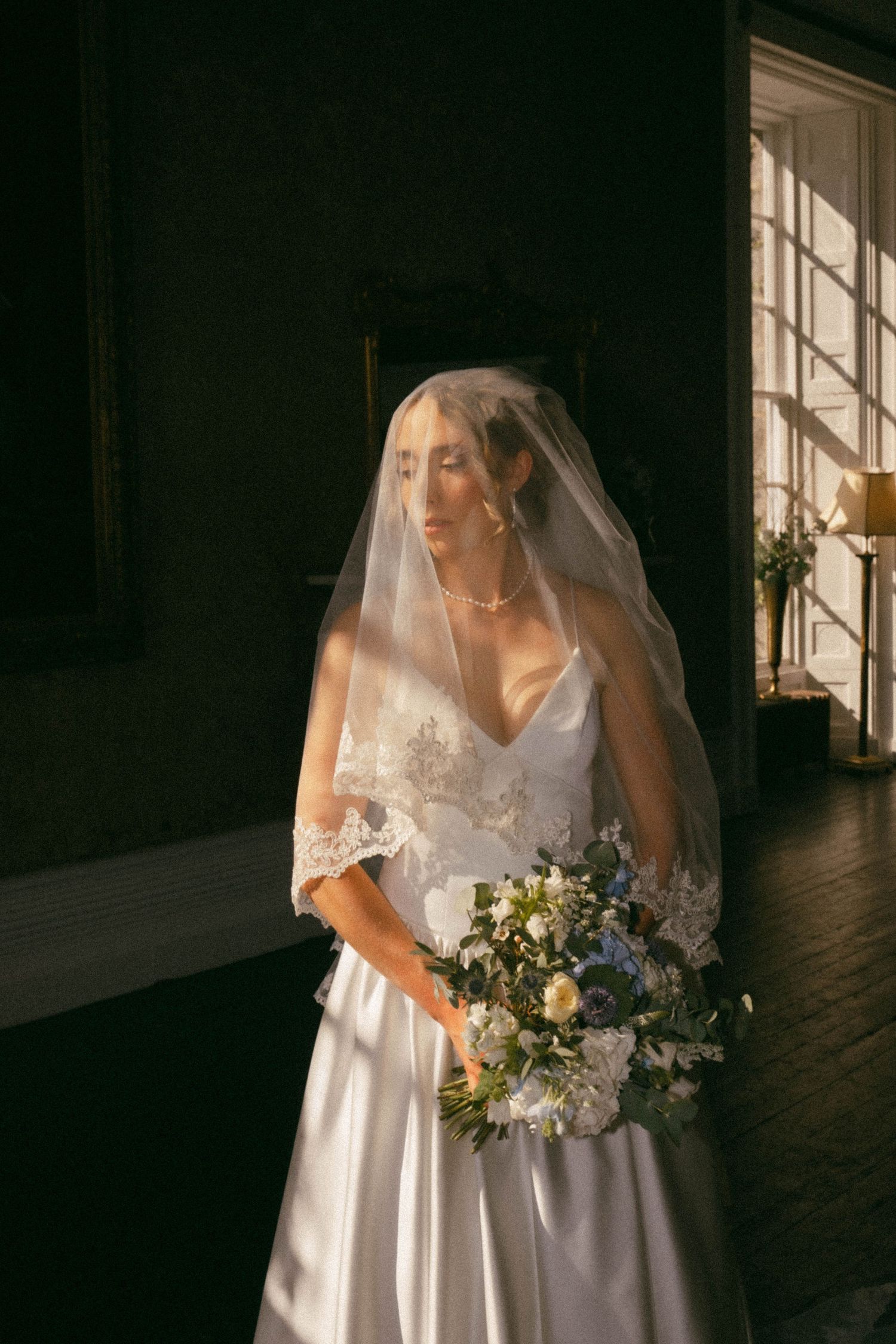 A bride in a white wedding dress and veil holding a bouquet stands in front of a window with natural light streaming in.