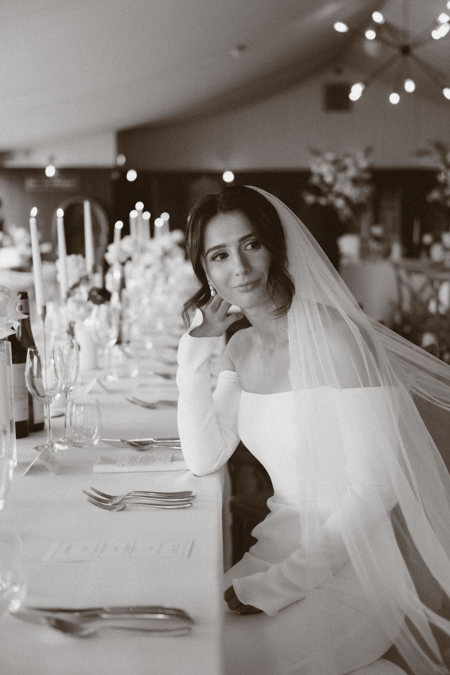 Bride in white gown and veil sits at elegantly set wedding reception table with candles and florals, black and white photo.