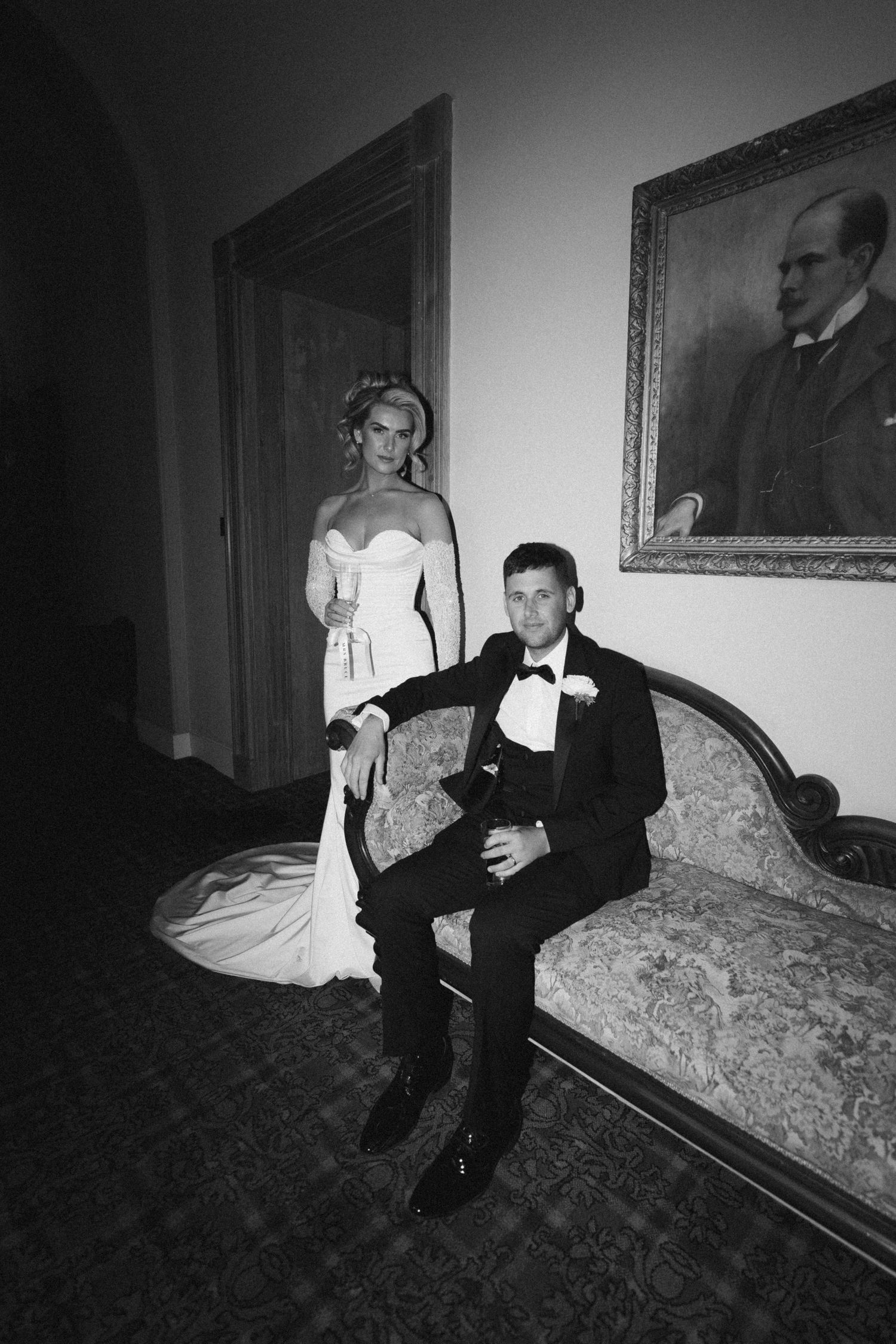 Black and white wedding photo showing couple posed formally on antique sofa with portrait on wall.