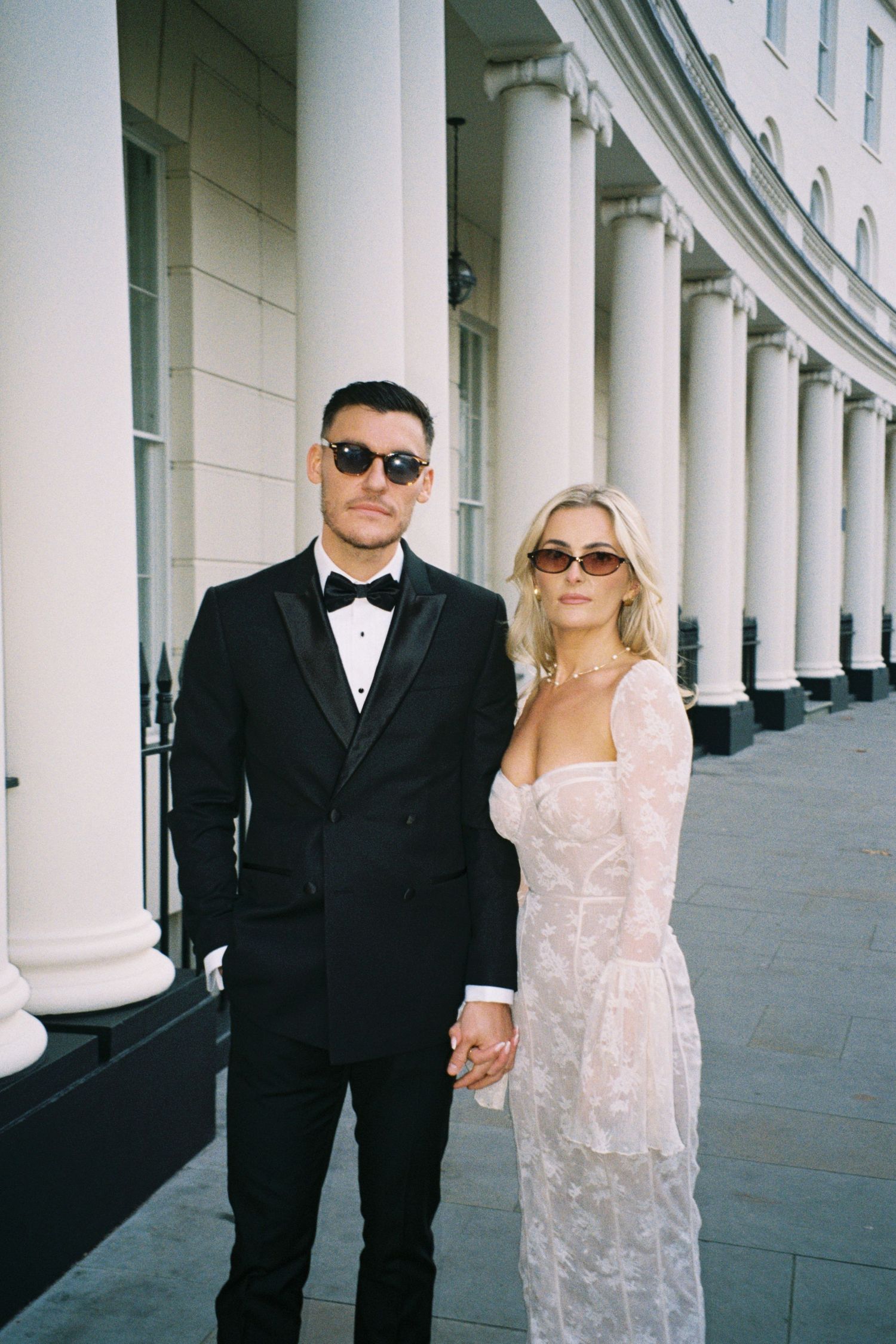 A couple in formal attire poses together in front of white columns, wearing sunglasses and holding hands.