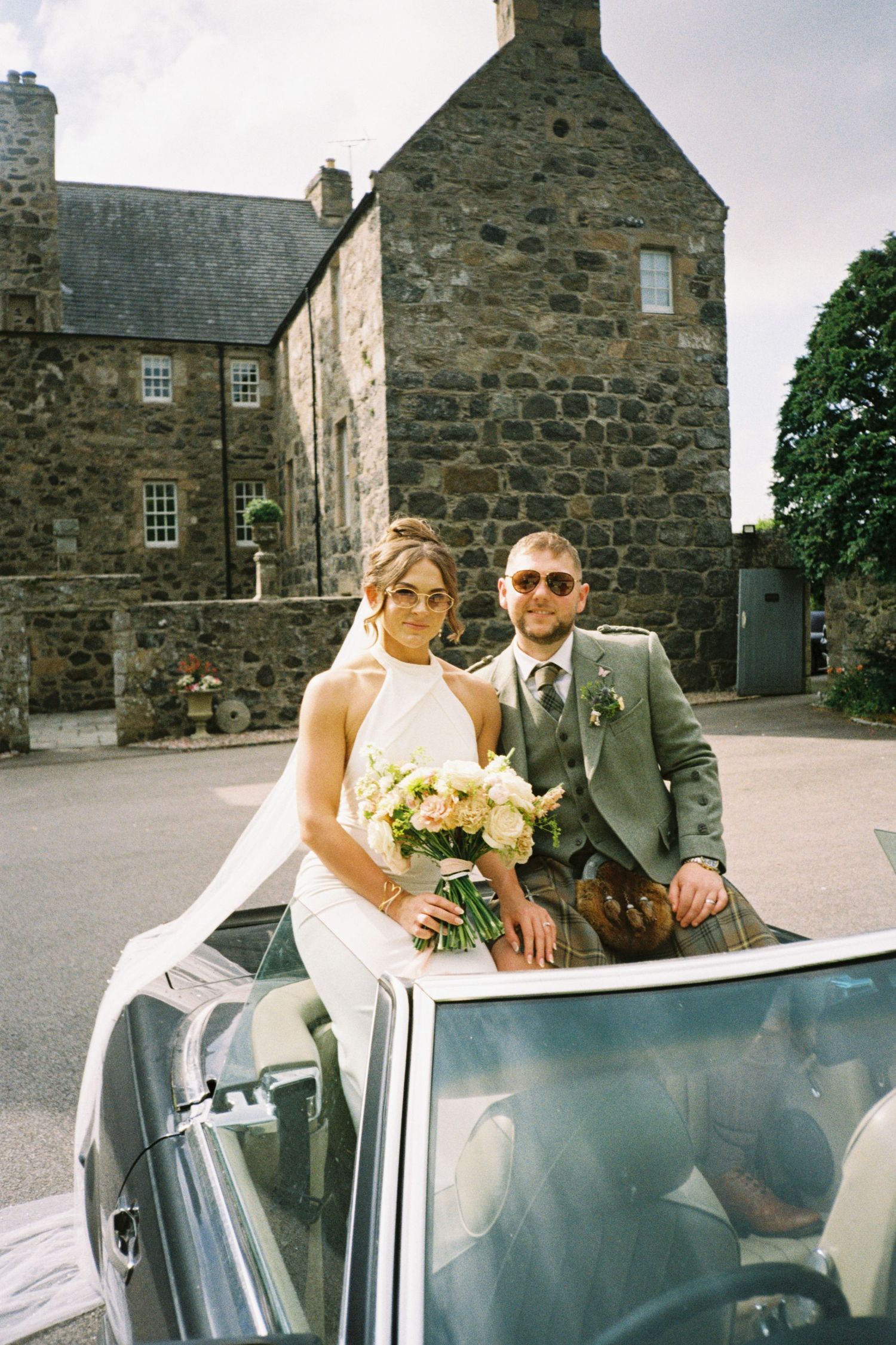 A wedding couple poses with their vintage car in front of a rustic stone building in Scotland.