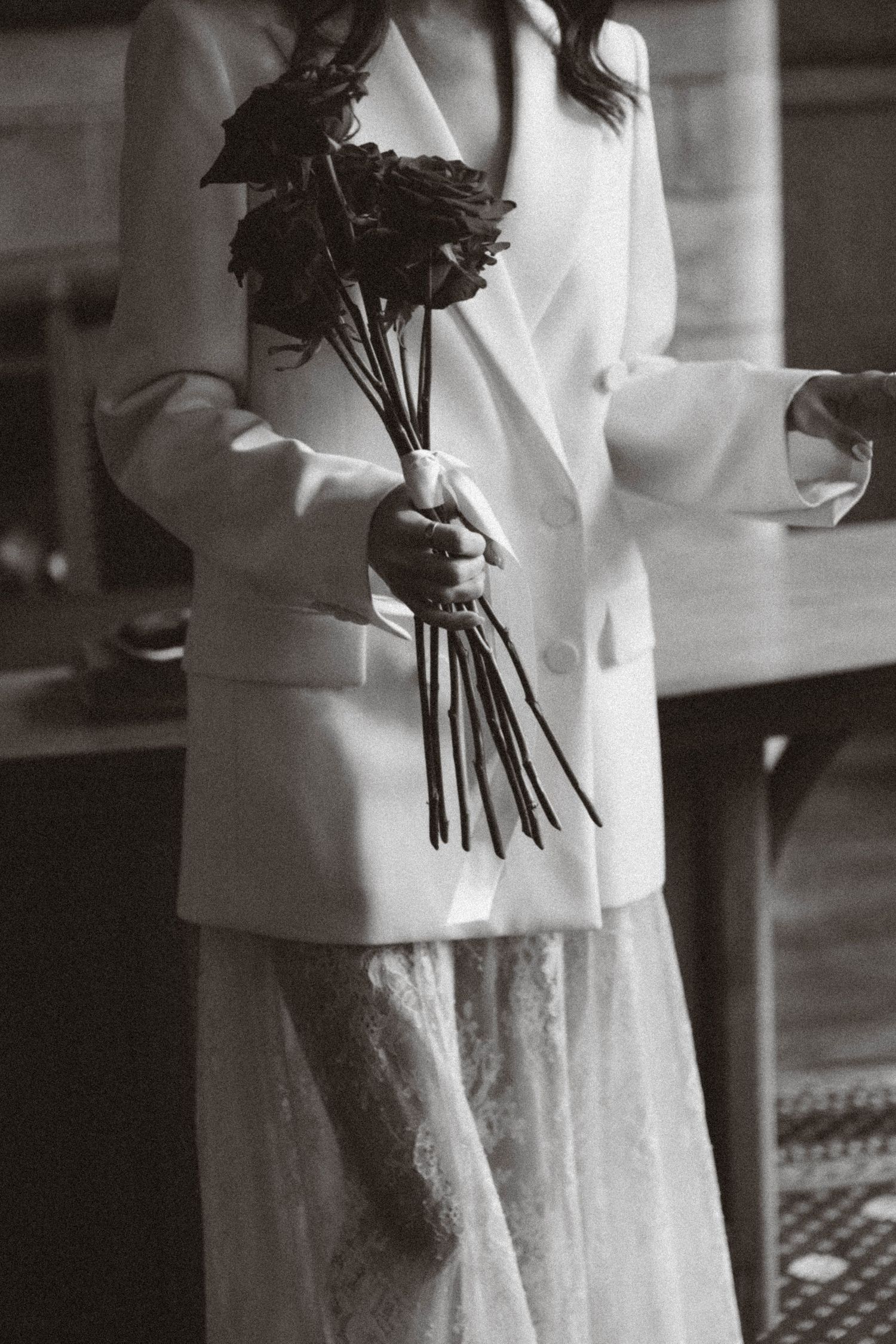 A black and white photo showing a person in a white blazer and lace dress holding a bouquet of dark roses.