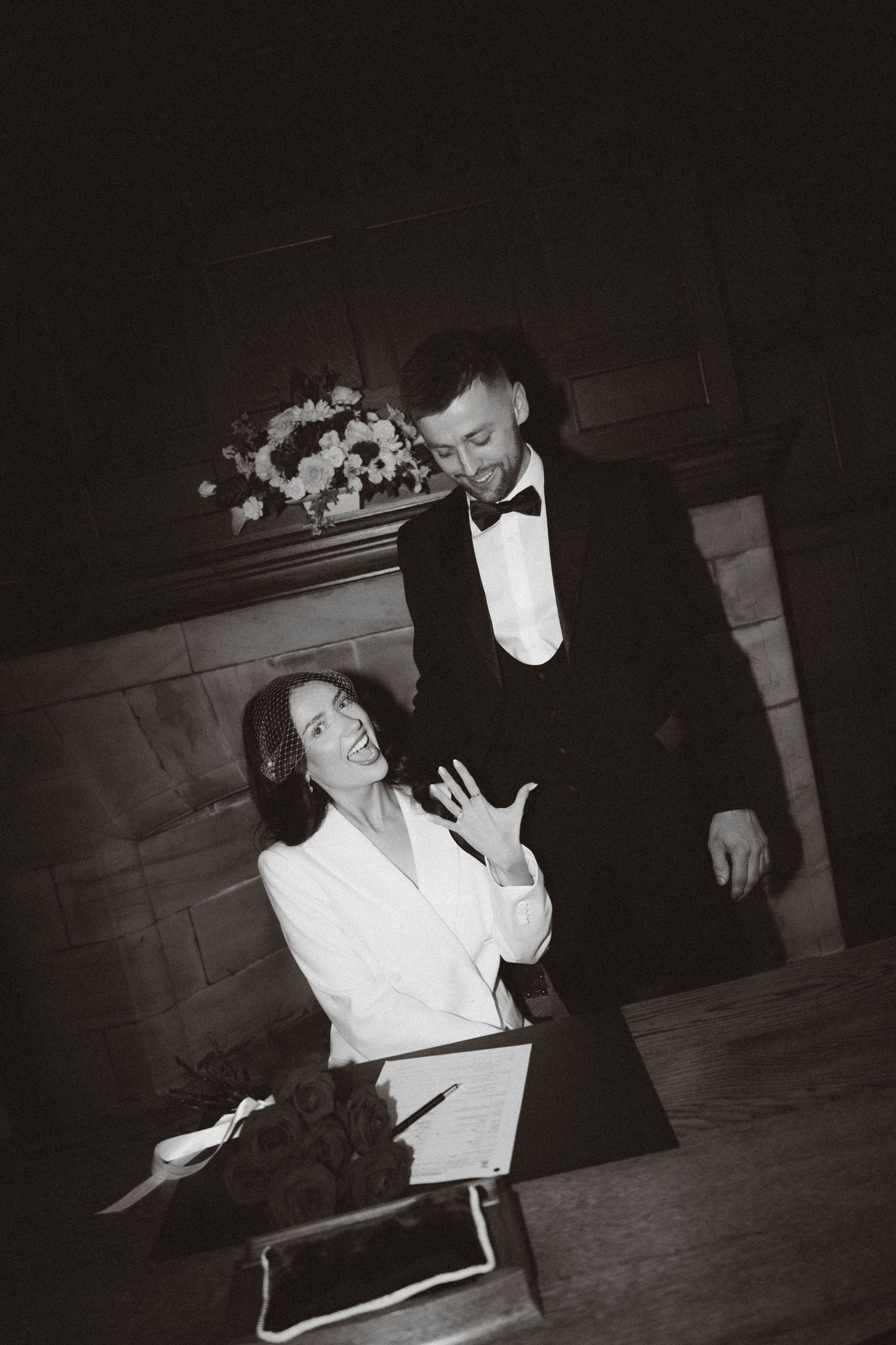 A black and white photograph shows two people in formal attire engaged in an animated conversation at an event.