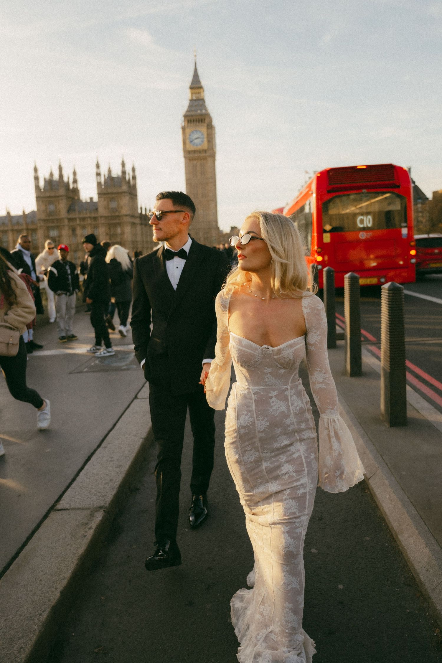 A bride in a white lace dress walks across Westminster Bridge in London with Big Ben and red buses in the background.