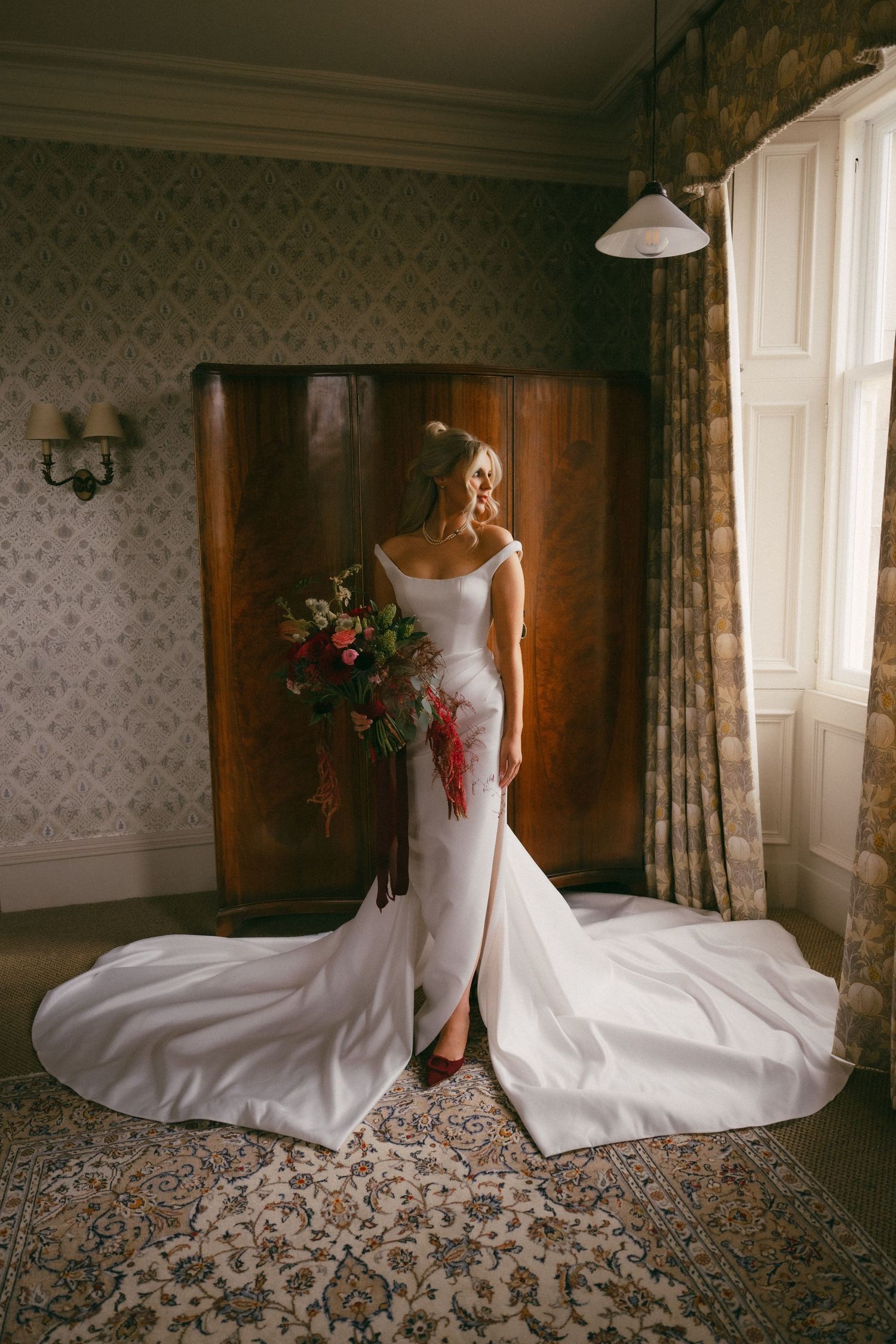 A stunning white wedding gown with a long train and floral bouquet photographed in an elegant room with patterned wallpaper.