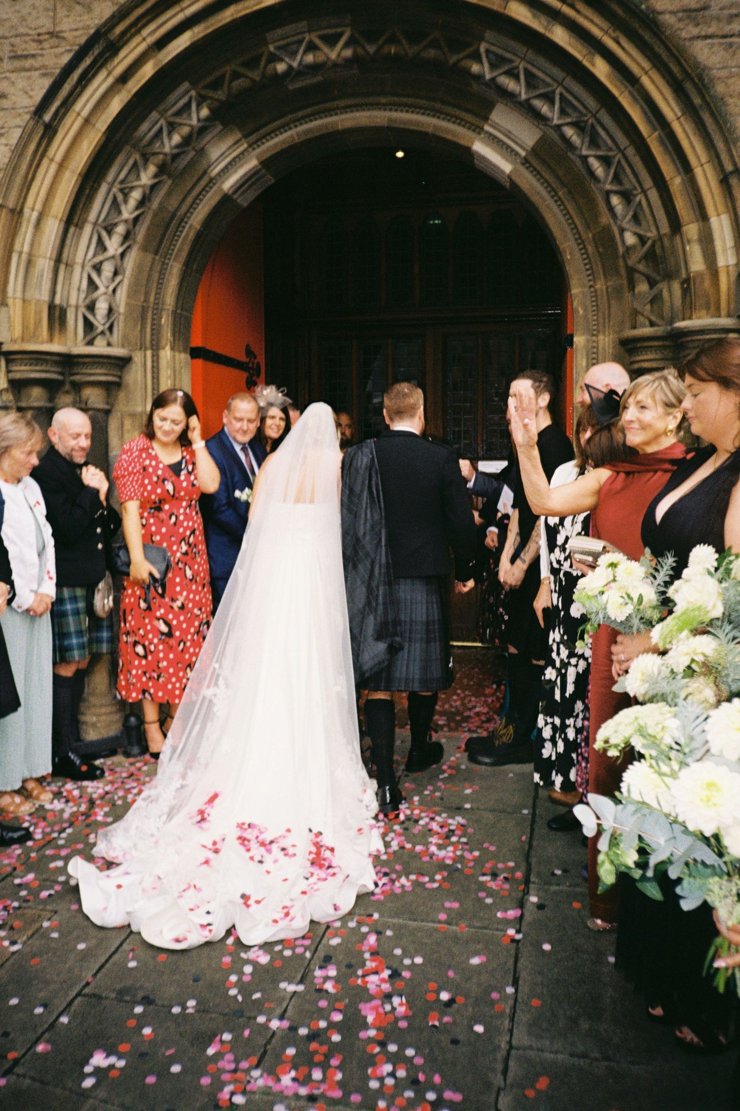 Wedding guests celebrate with rose petals as newlyweds exit through a grand stone archway at a historic venue.
