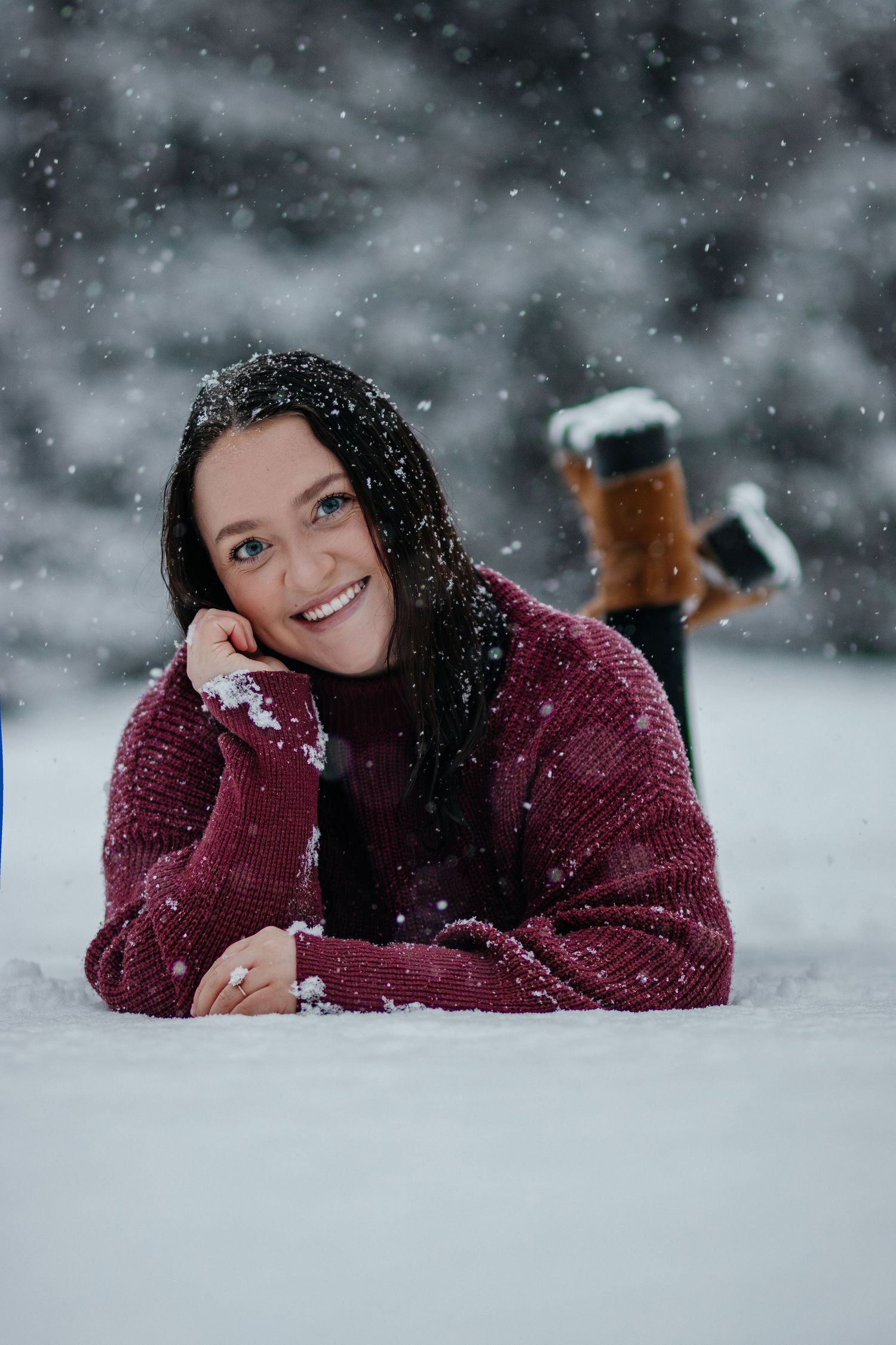 Person in burgundy sweater lying in snow with snowflakes falling during winter photoshoot.